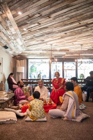People gather in a rustic space with wooden ceiling beams and floor cushions for a relaxed community gathering.