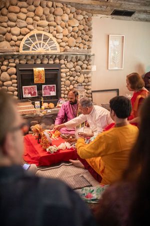 A traditional Hindu ceremony takes place in front of a stone fireplace with participants seated on red fabric.
