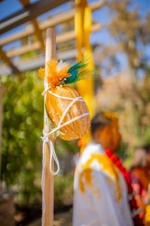 Decorative orange pumpkin ornament with green feathers and white ribbon hanging against blurred autumn background.