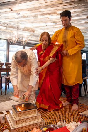 People in traditional red and yellow attire perform a ceremonial ritual at a wooden table with decorative objects.