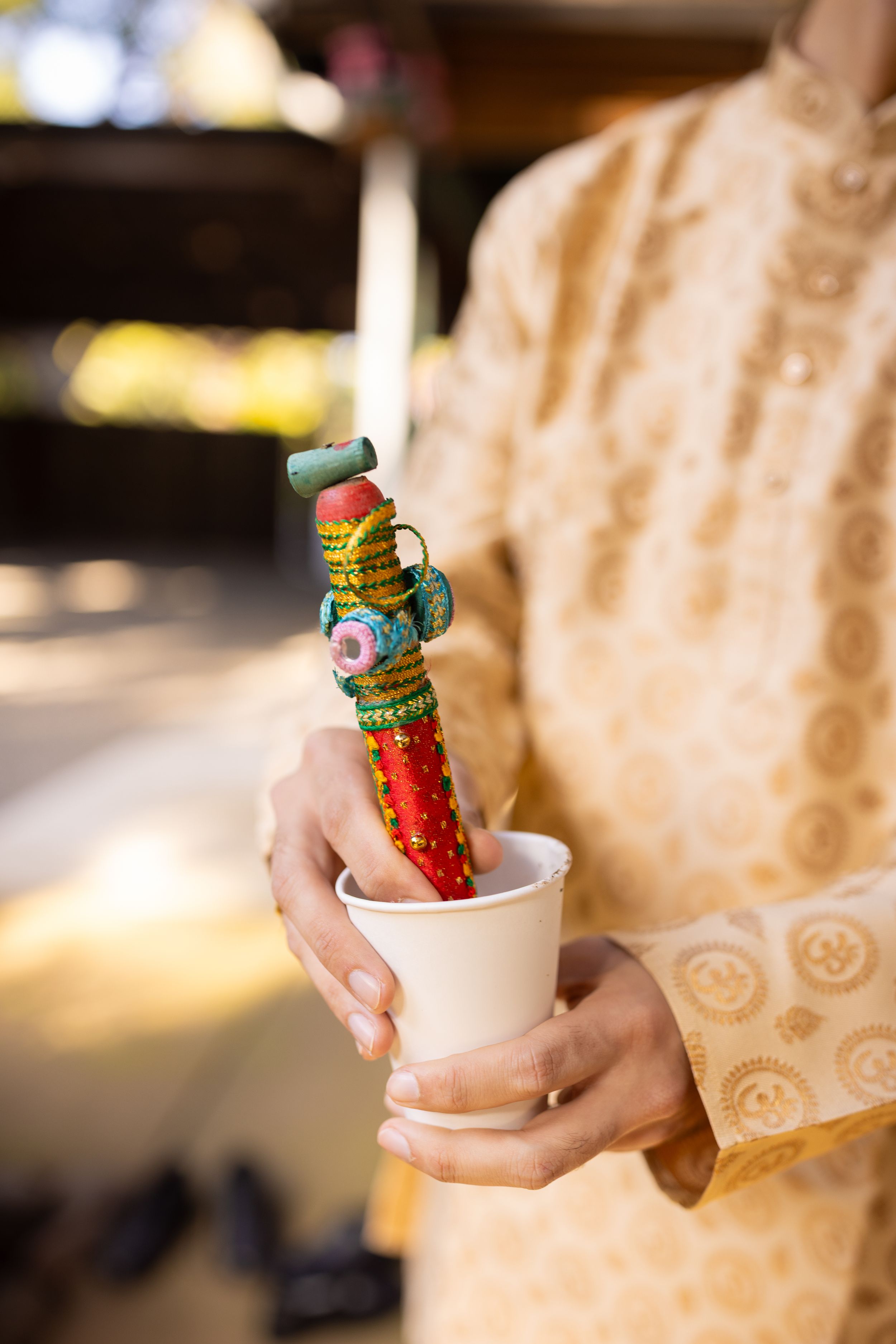 Hands holding a decorative Indian wedding ritual item with colorful beads and embellishments in a small cup.