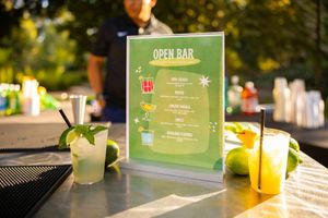 Colorful cocktails and menu cards displayed on a wooden bar counter with natural lighting and greenery.