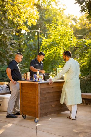 A mobile bar setup at an outdoor event with staff serving drinks in a natural sunlit garden setting.