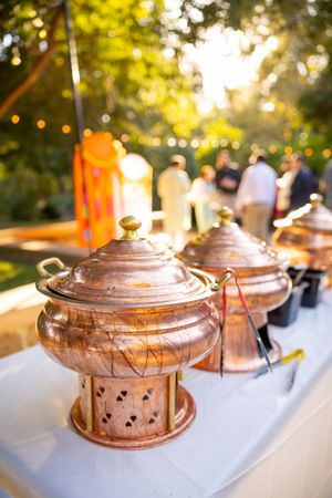 Copper chafing dishes lined up on a white tablecloth at an outdoor catering event during golden hour.
