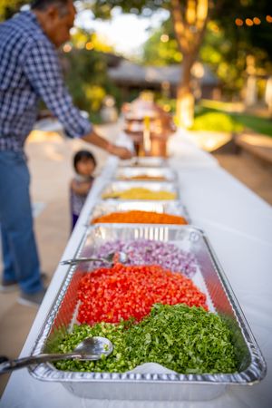 Buffet line of fresh chopped vegetables including tomatoes, onions, and leafy greens in metal serving trays.