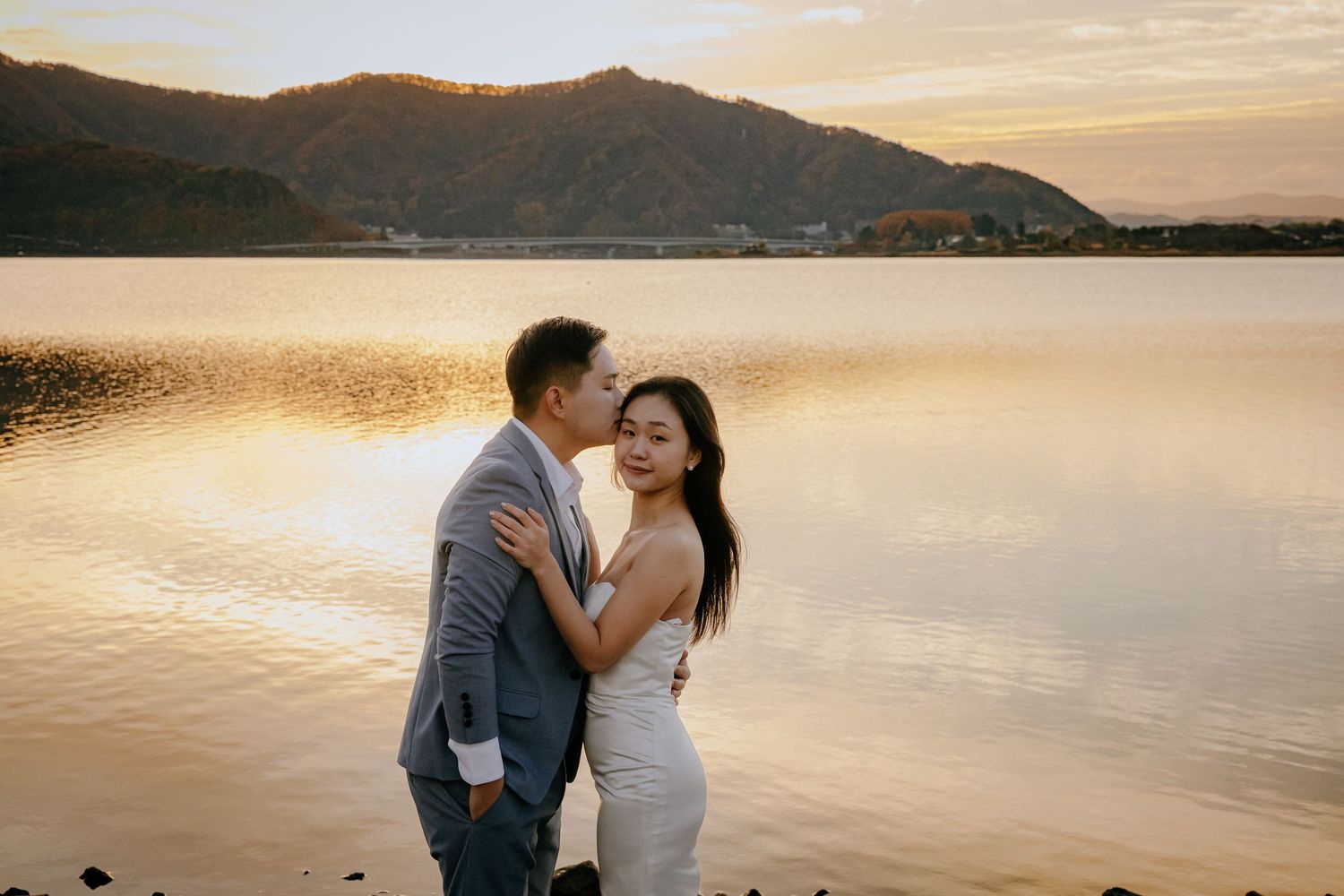 A couple embraces during sunset by a serene lake with mountains in the background.