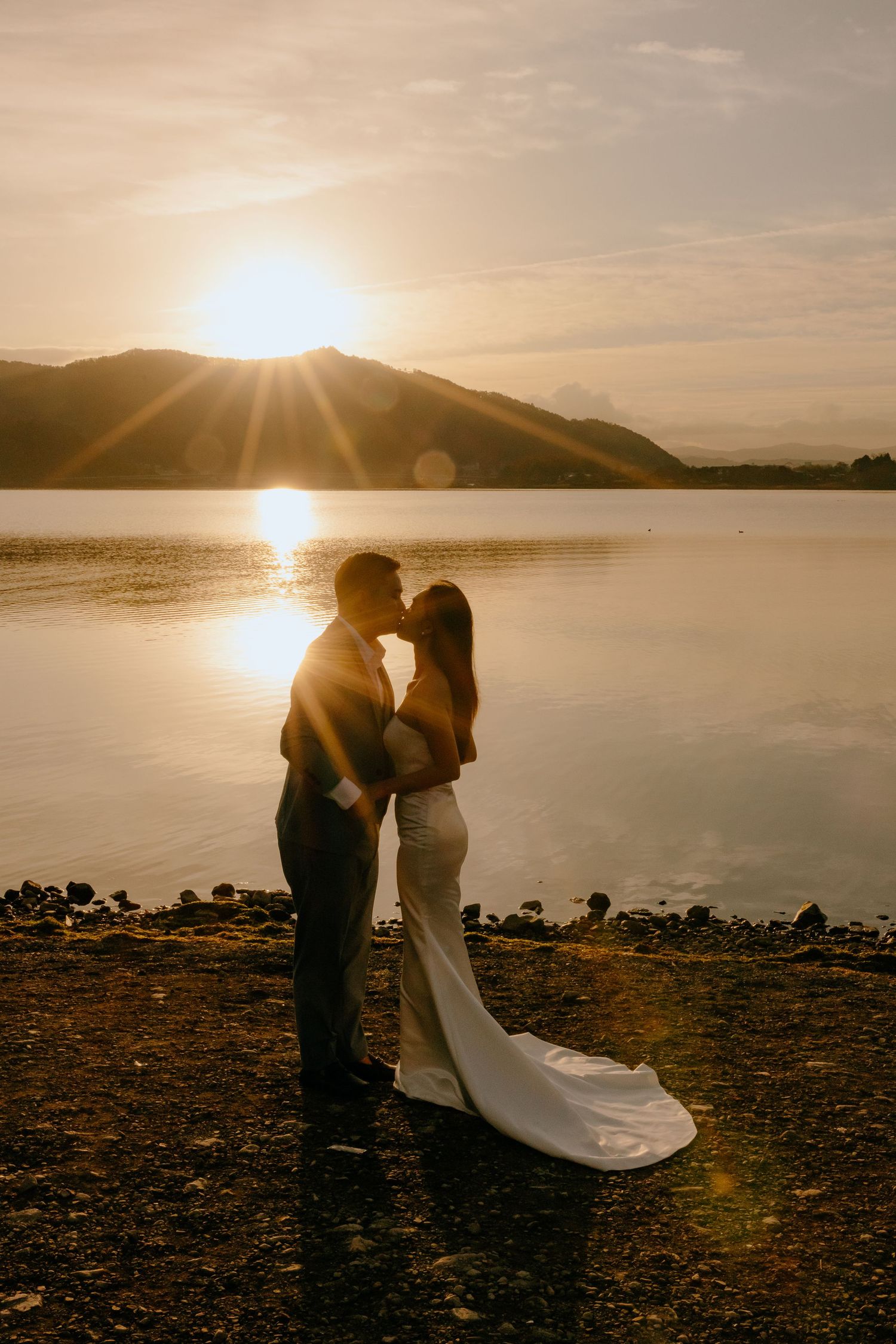 A romantic silhouette of a couple embracing by a serene lake at sunset with mountains in the background.