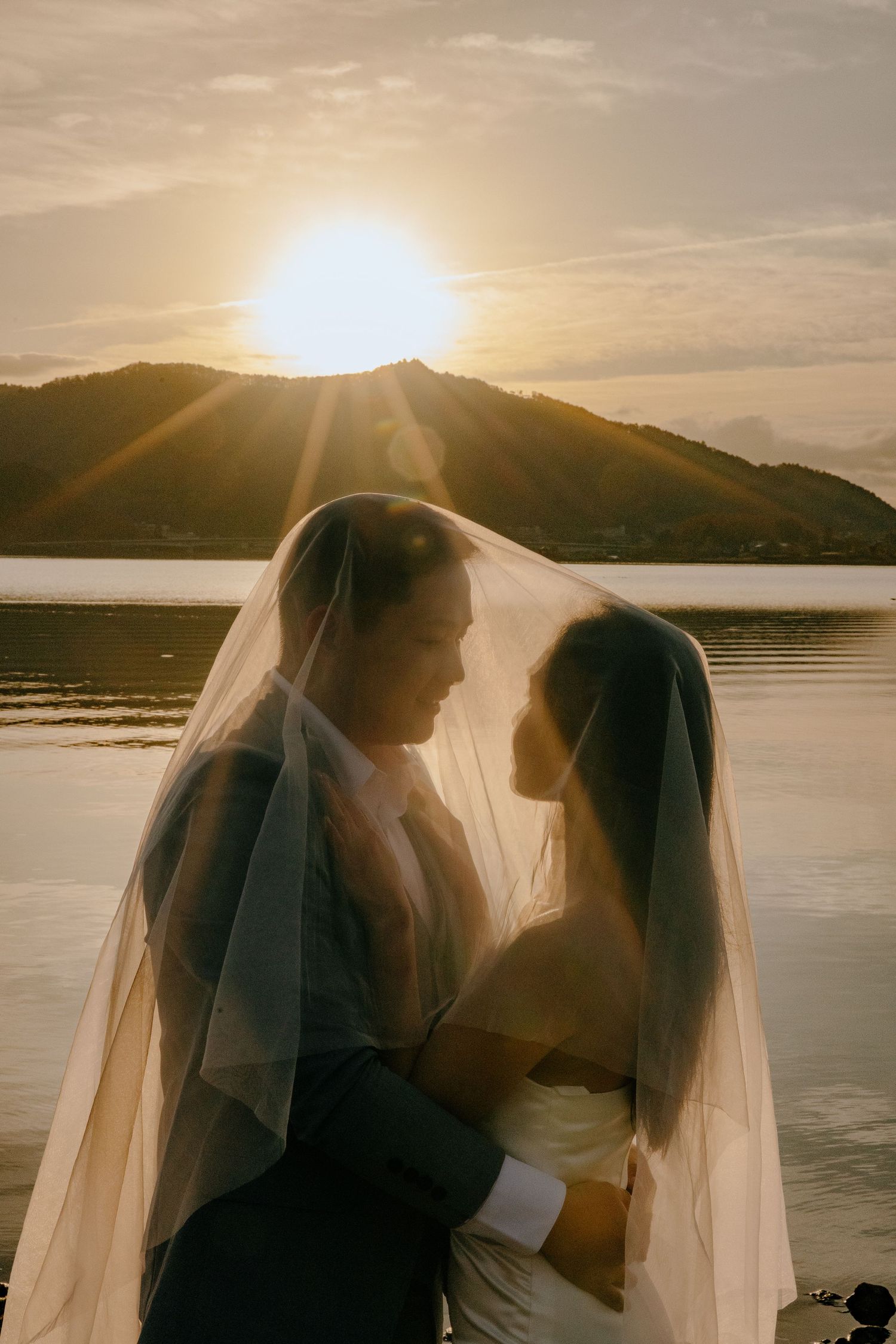 Silhouetted couple in wedding attire share intimate moments at sunset by a serene lake with mountains in the background.