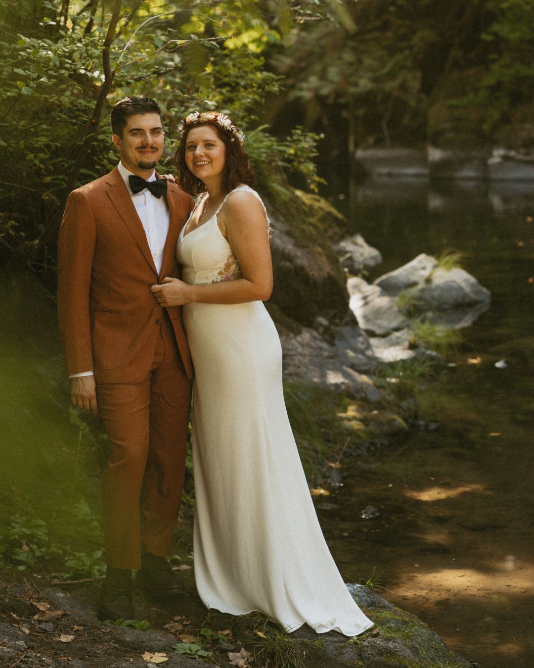 Newlyweds pose in a rustic woodland setting with a stream in the background.