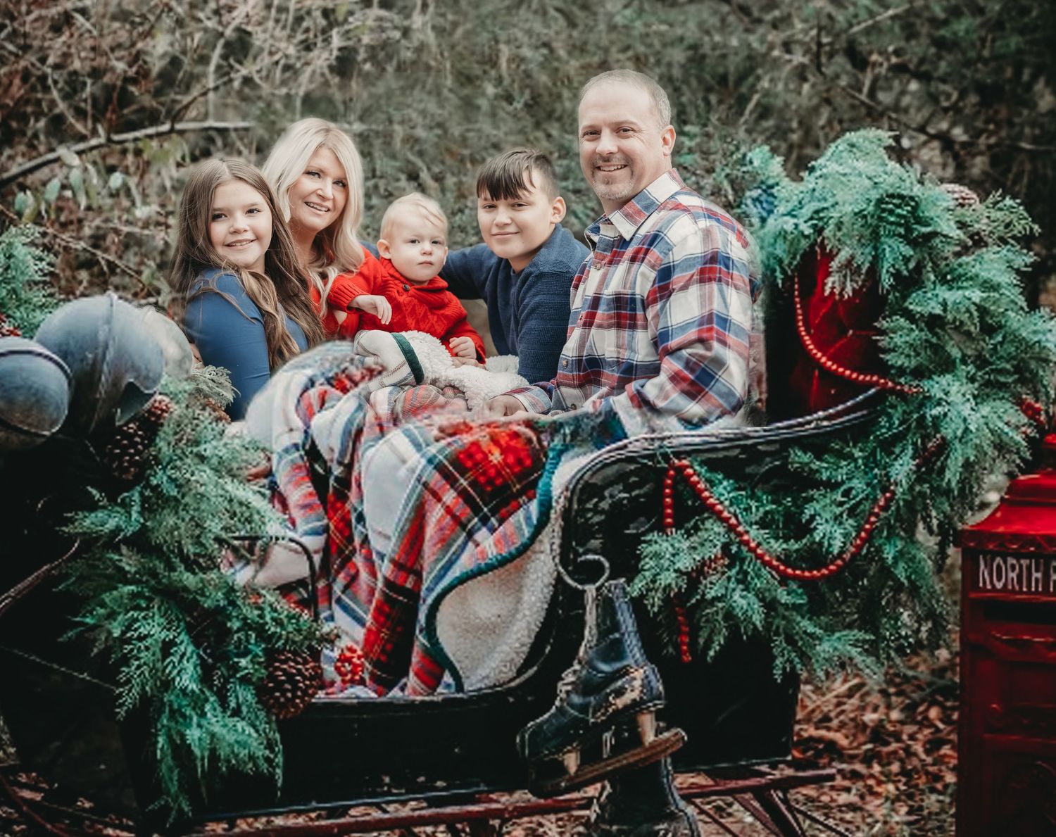 Family holiday photo in a rustic sleigh surrounded by Christmas greenery and plaid blankets.