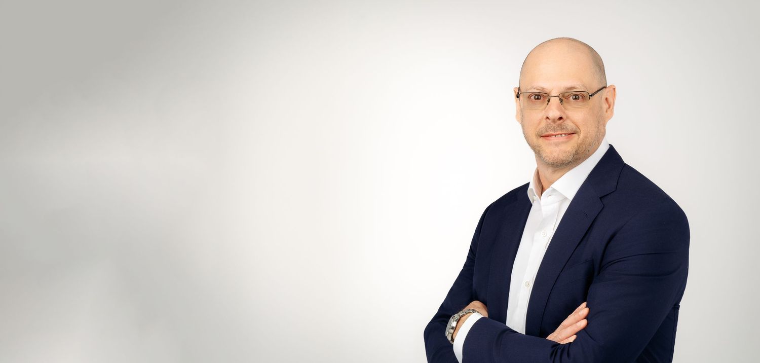 Professional portrait of someone in a navy blazer and white shirt with arms crossed against a light gray background.