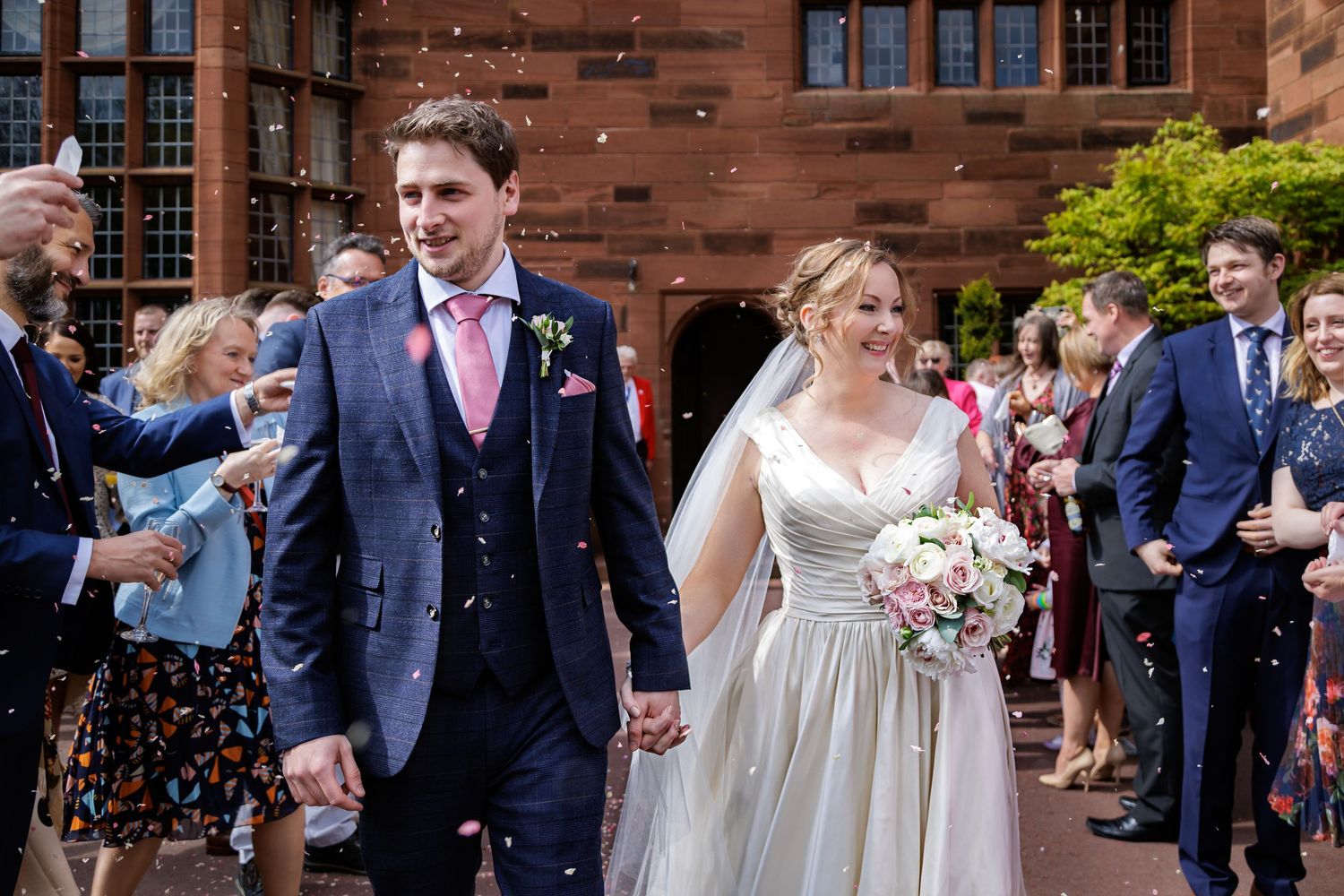 Wedding guests throw confetti at a couple outside a red brick building during a joyful celebration.
