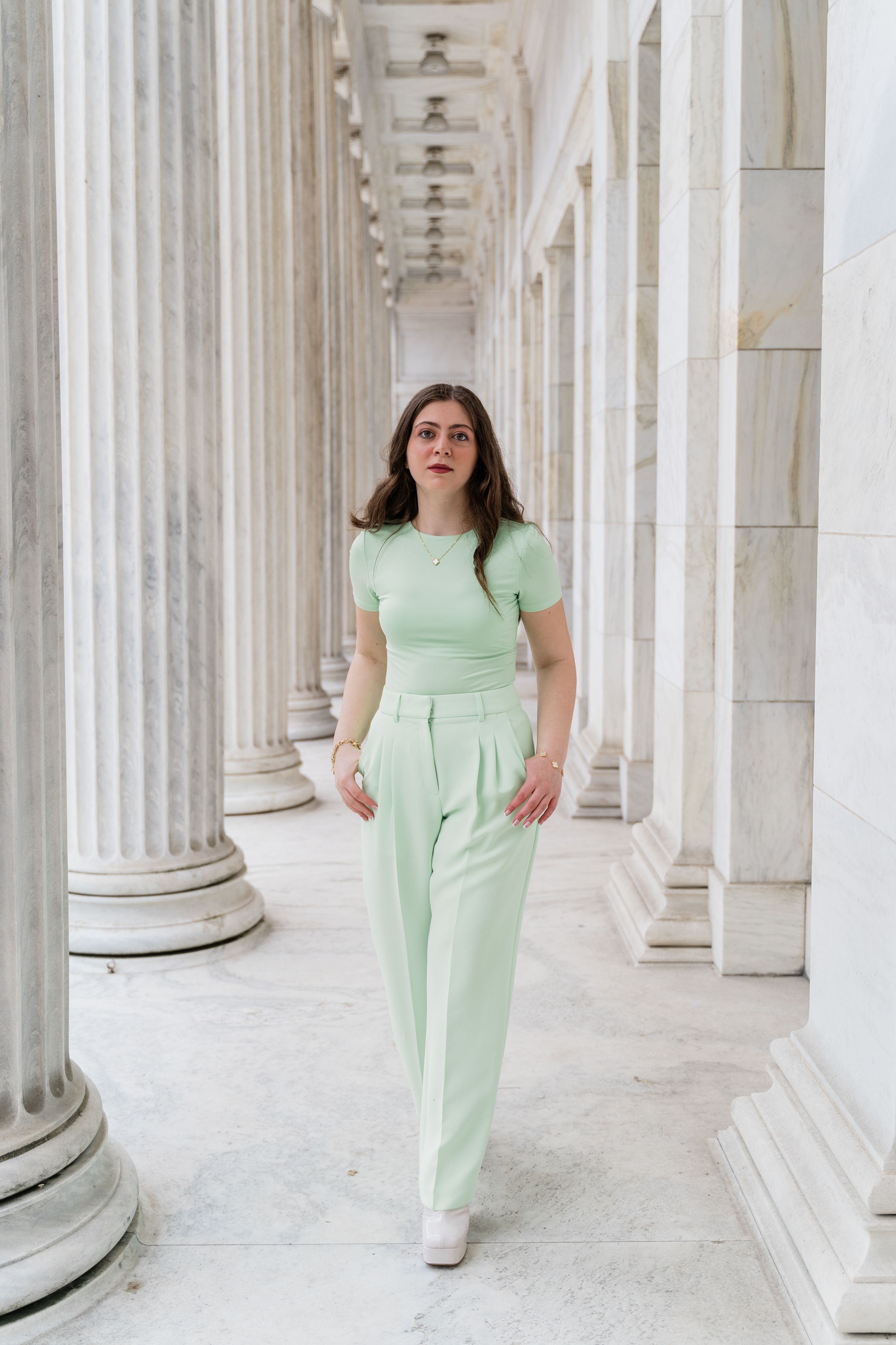 Woman in mint green jumpsuit poses elegantly between white marble columns in a classic architectural setting.
