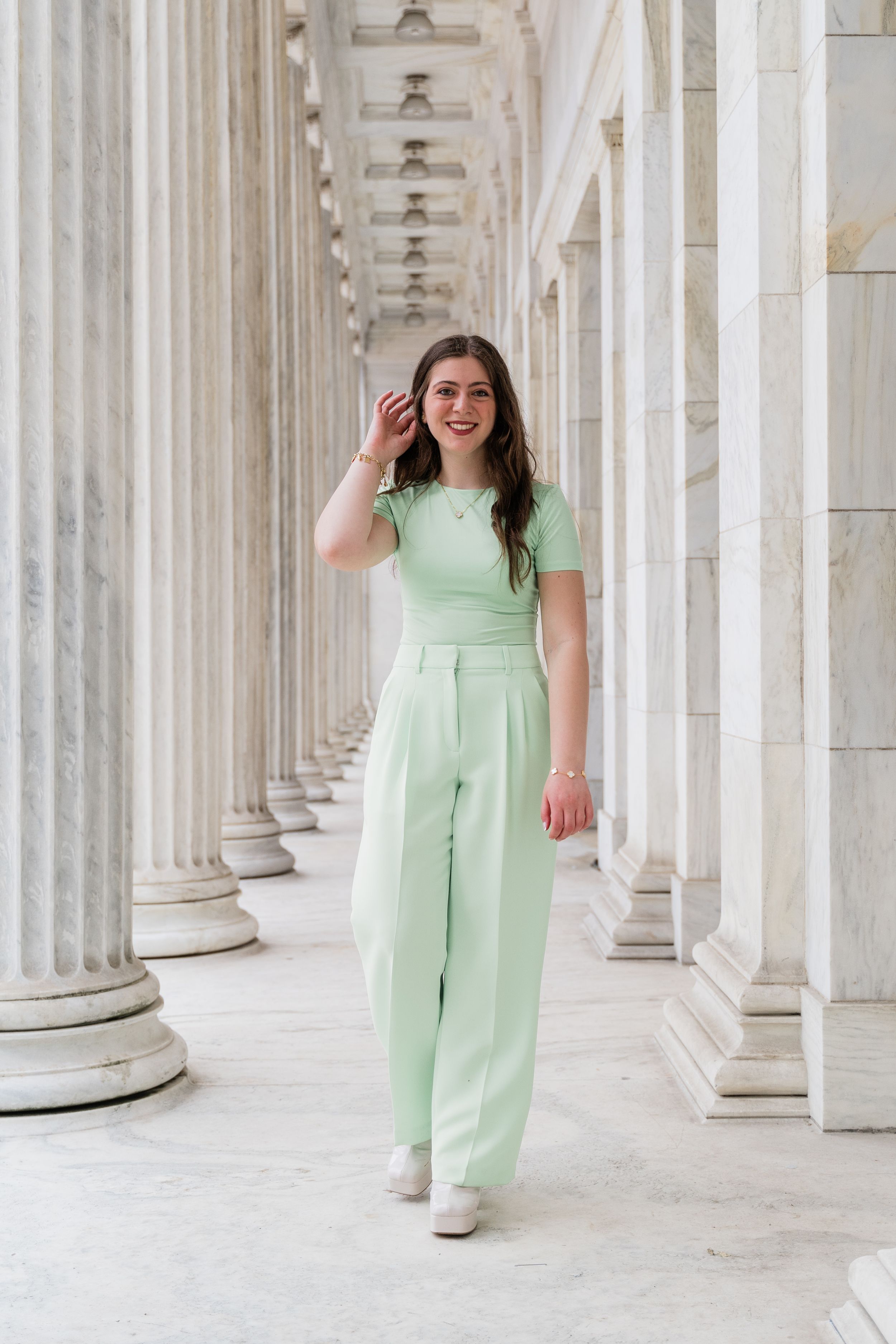 Person in mint green jumpsuit poses elegantly between classical white columns in architectural setting.