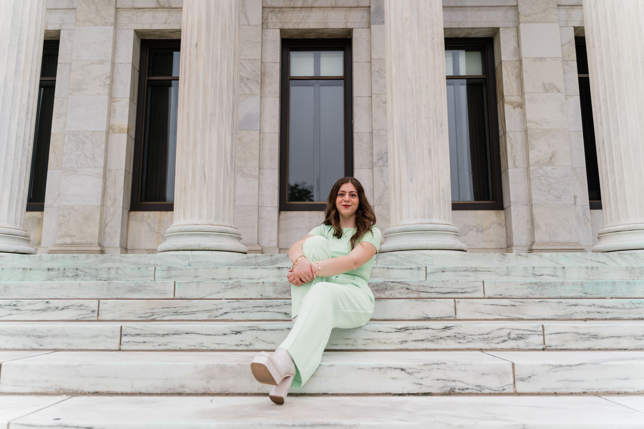 Someone in mint green pants sits casually on stone steps of a grand building with tall columns and windows.
