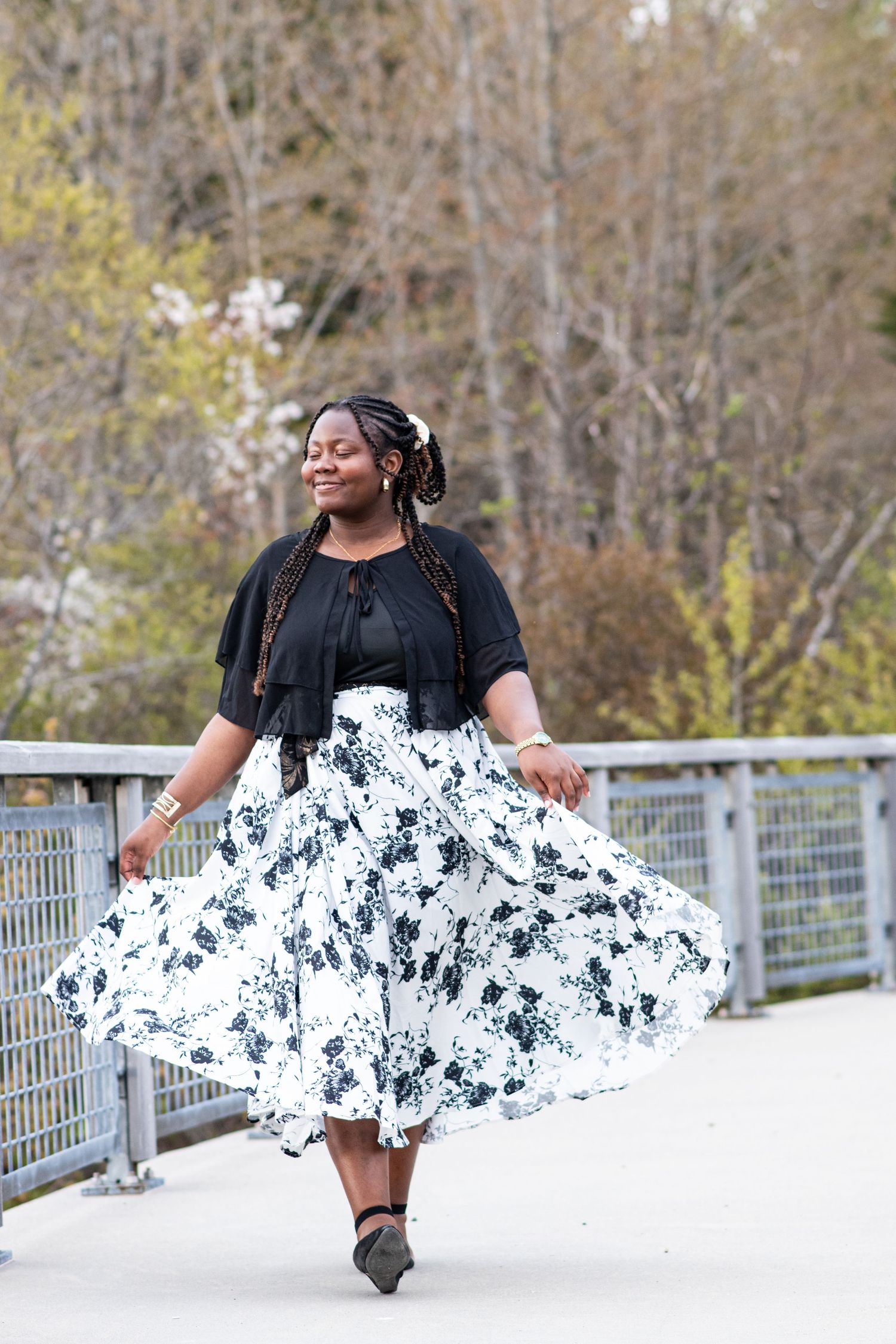 Joyful Senior Girl twirling in black and white floral skirt on bridge surrounded by spring trees. Photo by Xenophon Photography in Maine.