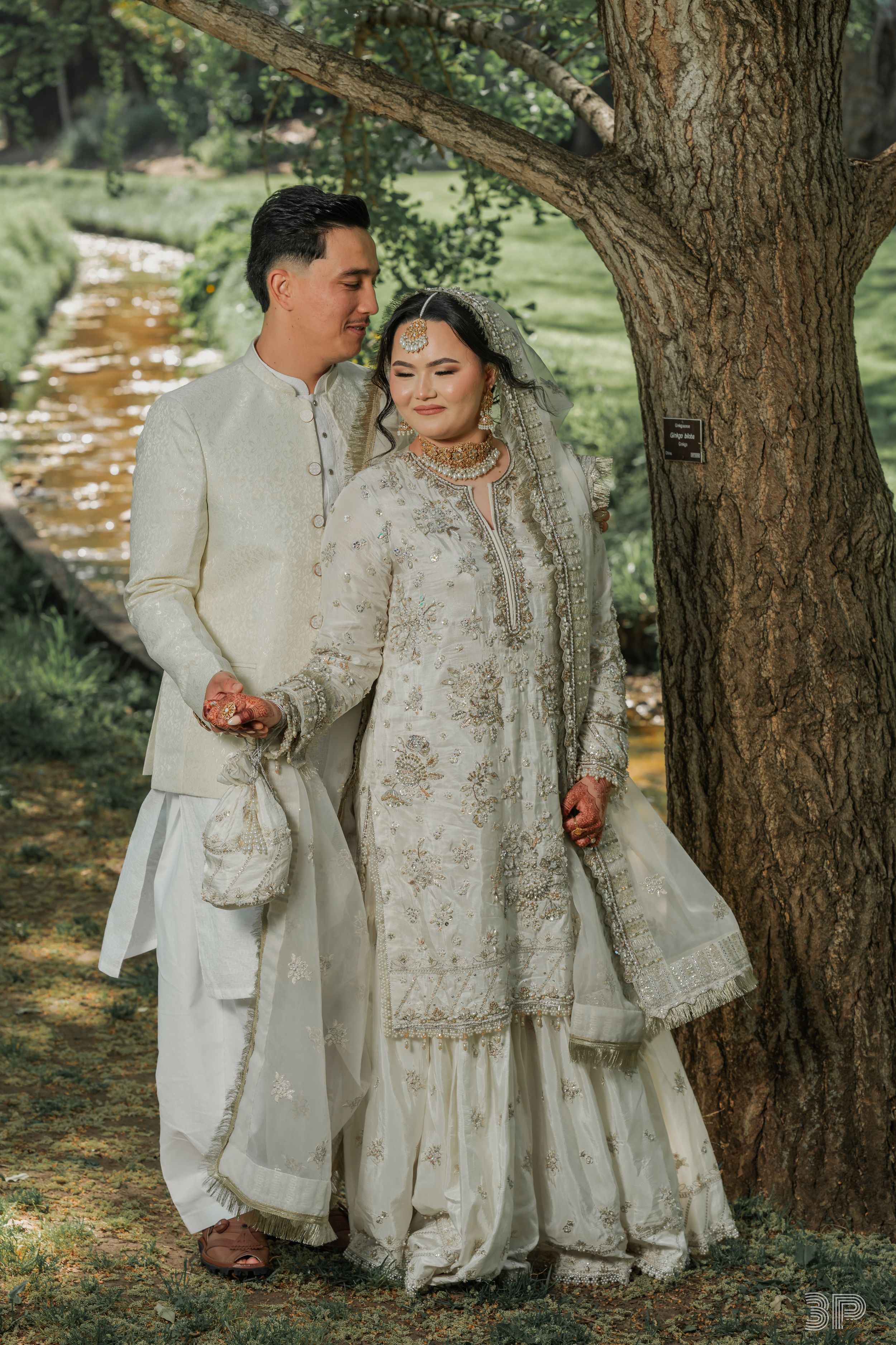 Couple in matching ornate white traditional wedding attire posing together by a tree.