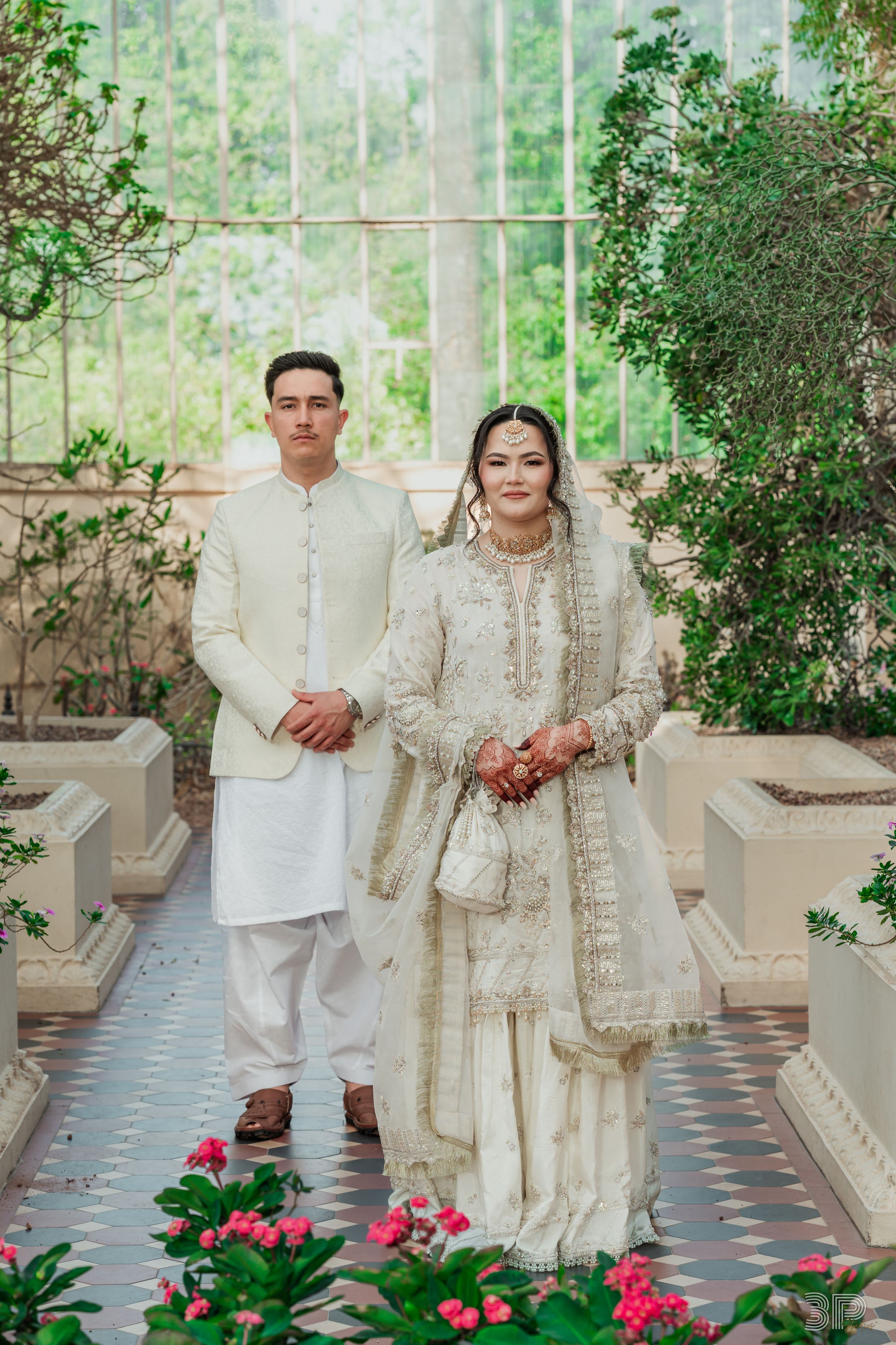 Wedding couple in elegant white outfits standing in a garden with pink flowers.