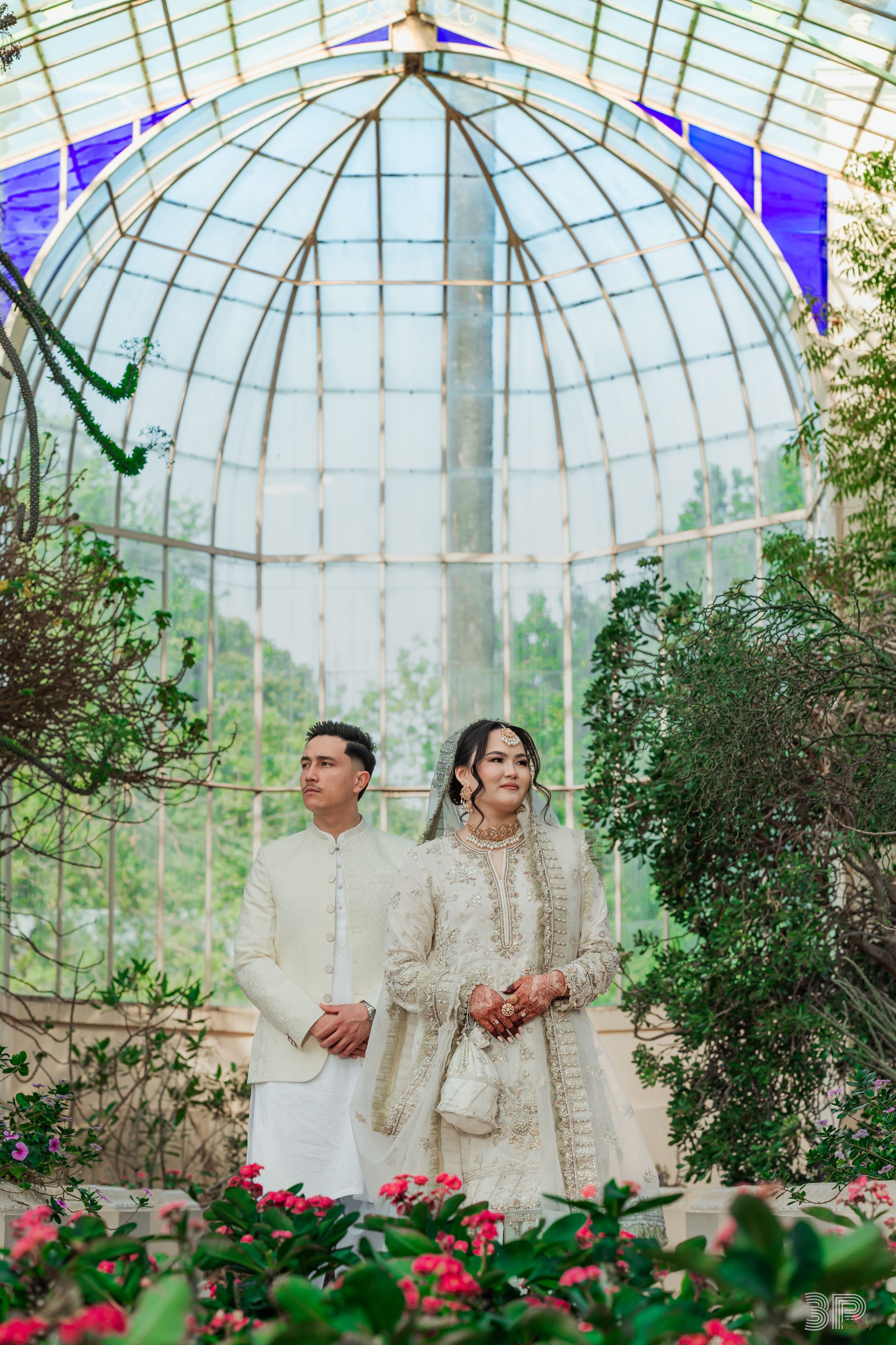 Couple in traditional white attire photographed under a glass dome conservatory with pink flowers.