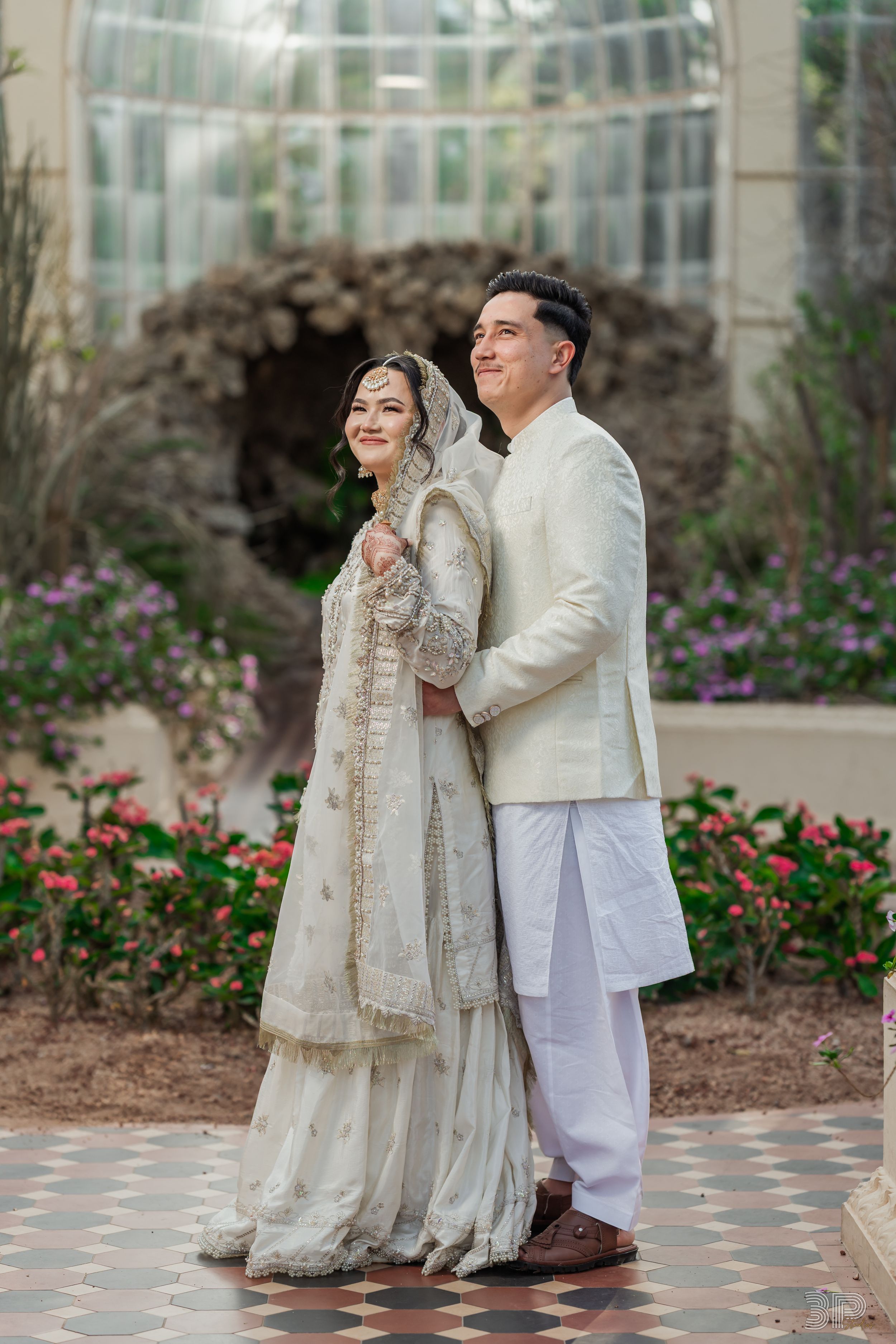 Couple in elegant white and cream outfits posing together in a garden setting with colorful flowers.