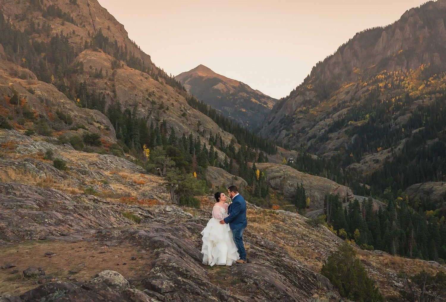 A couple in wedding attire stands on a rocky overlook surrounded by majestic mountain peaks at sunset.