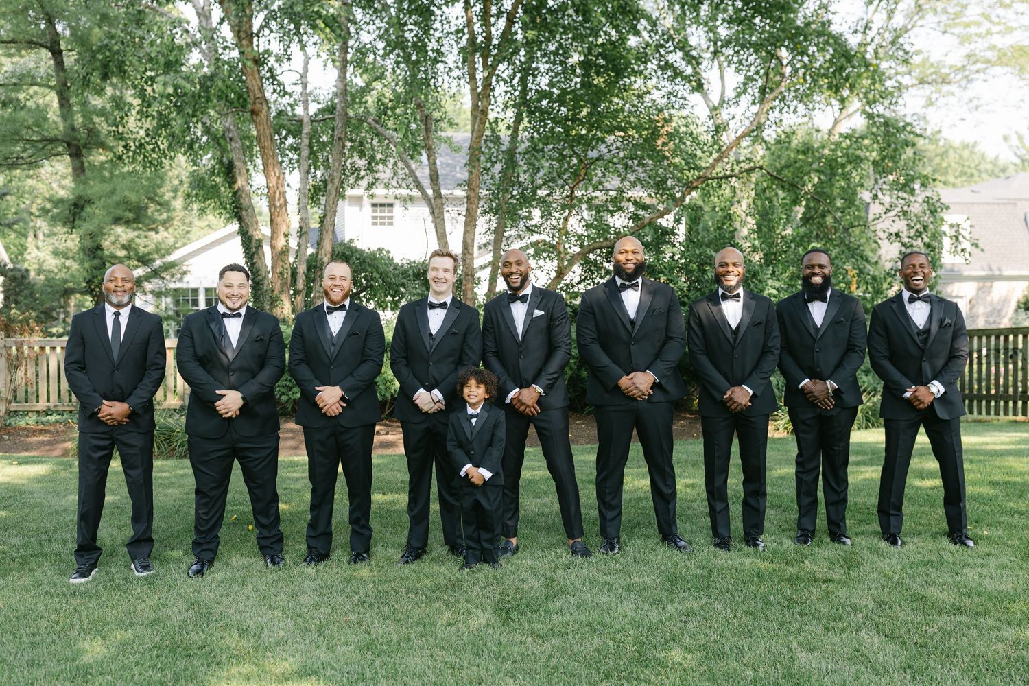Groomsmen in black tuxedos and bow ties pose together on a lush green lawn for formal wedding photos.