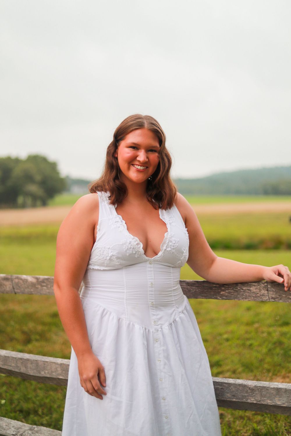 Woman in white dress smiling while leaning on wooden fence in countryside setting.