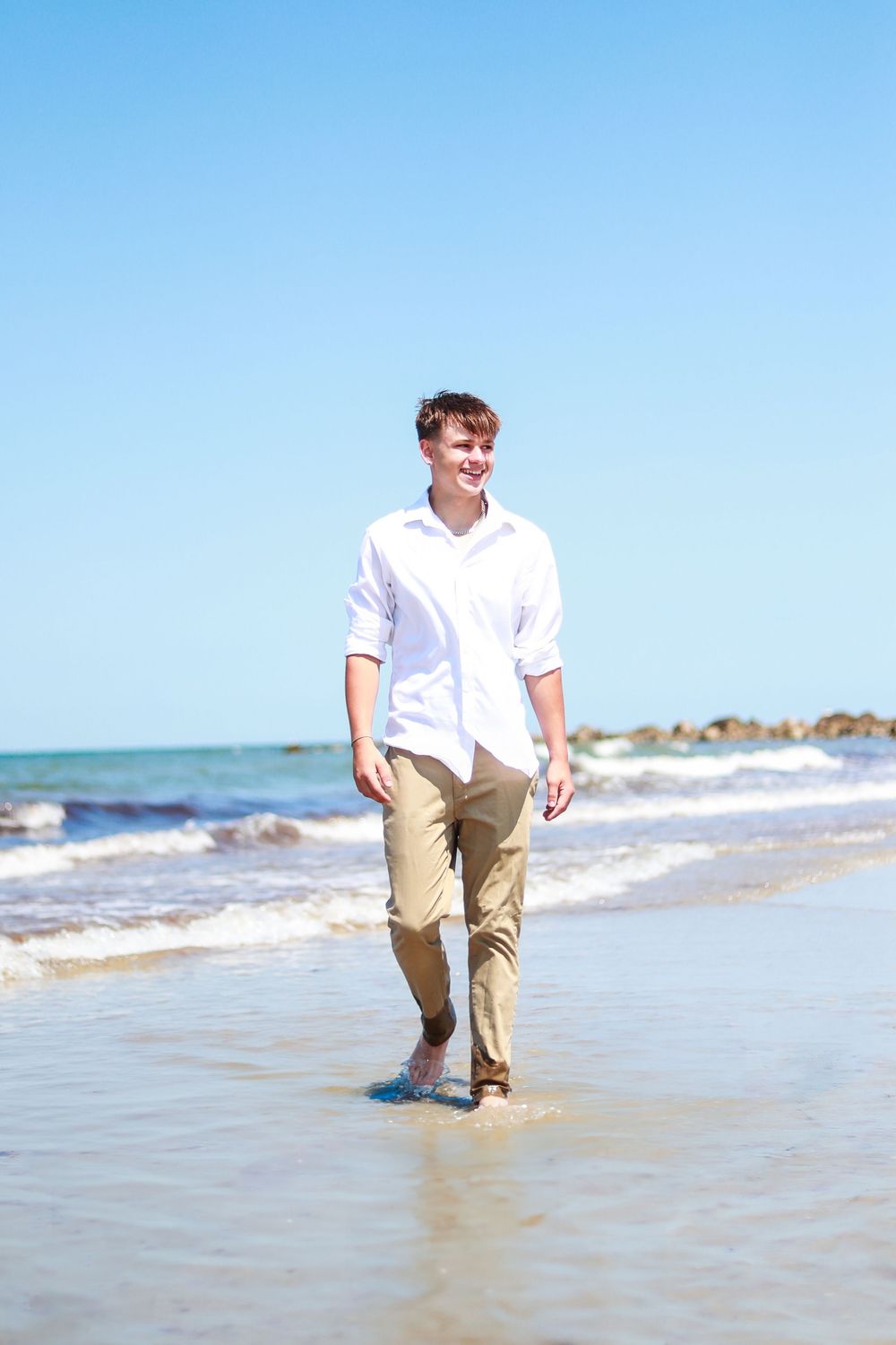 Person in white shirt and khaki pants walks along ocean shoreline.