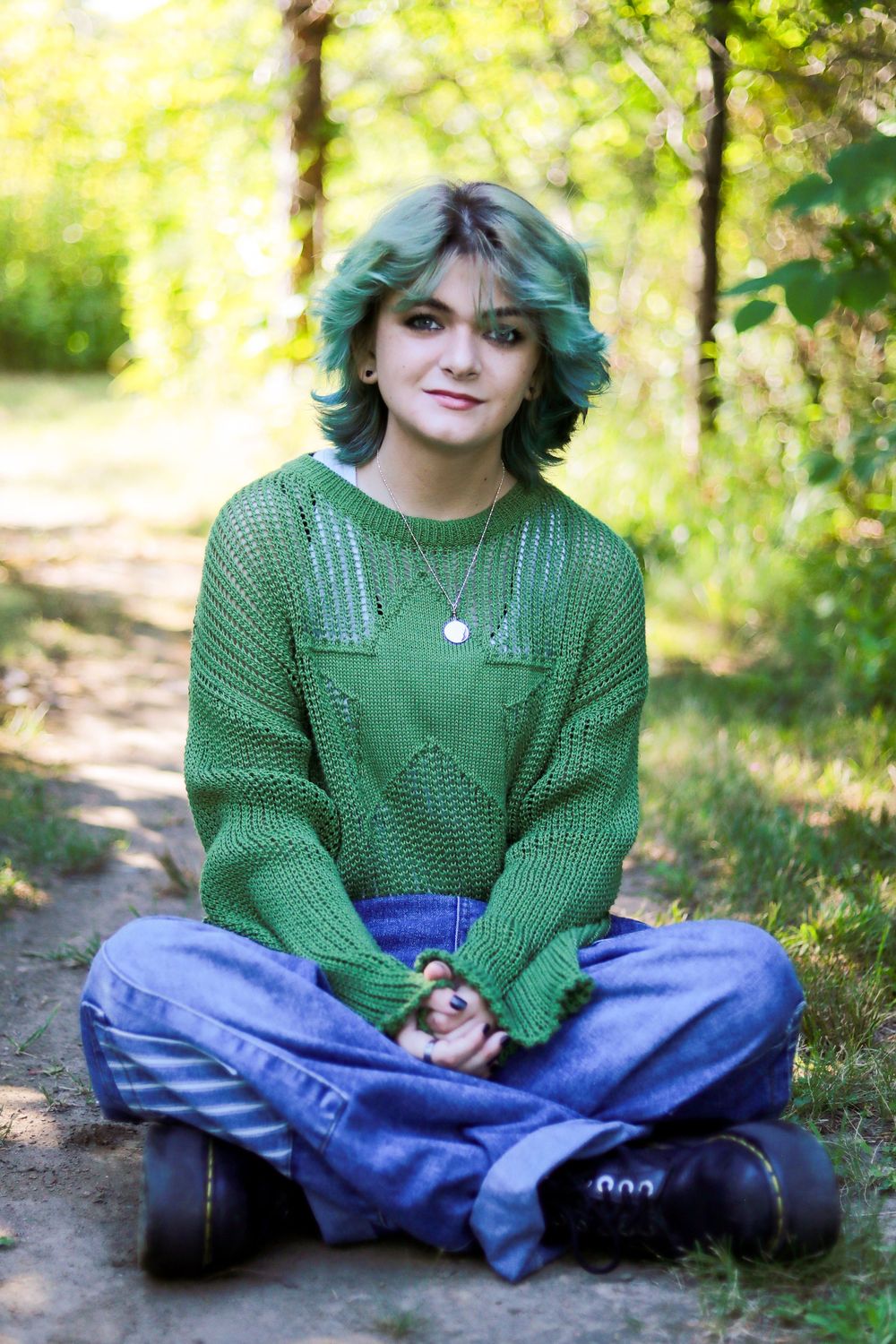 Person wearing green sweater sits cross-legged outdoors in natural setting.