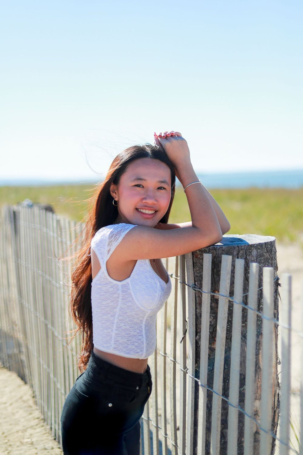 Person in white top poses playfully by wooden beach fence on sunny day.
