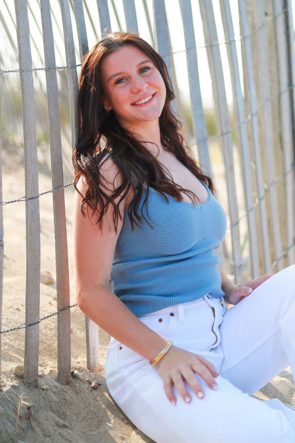 Portrait series against beach fence wearing blue top and white pants on sandy dunes.