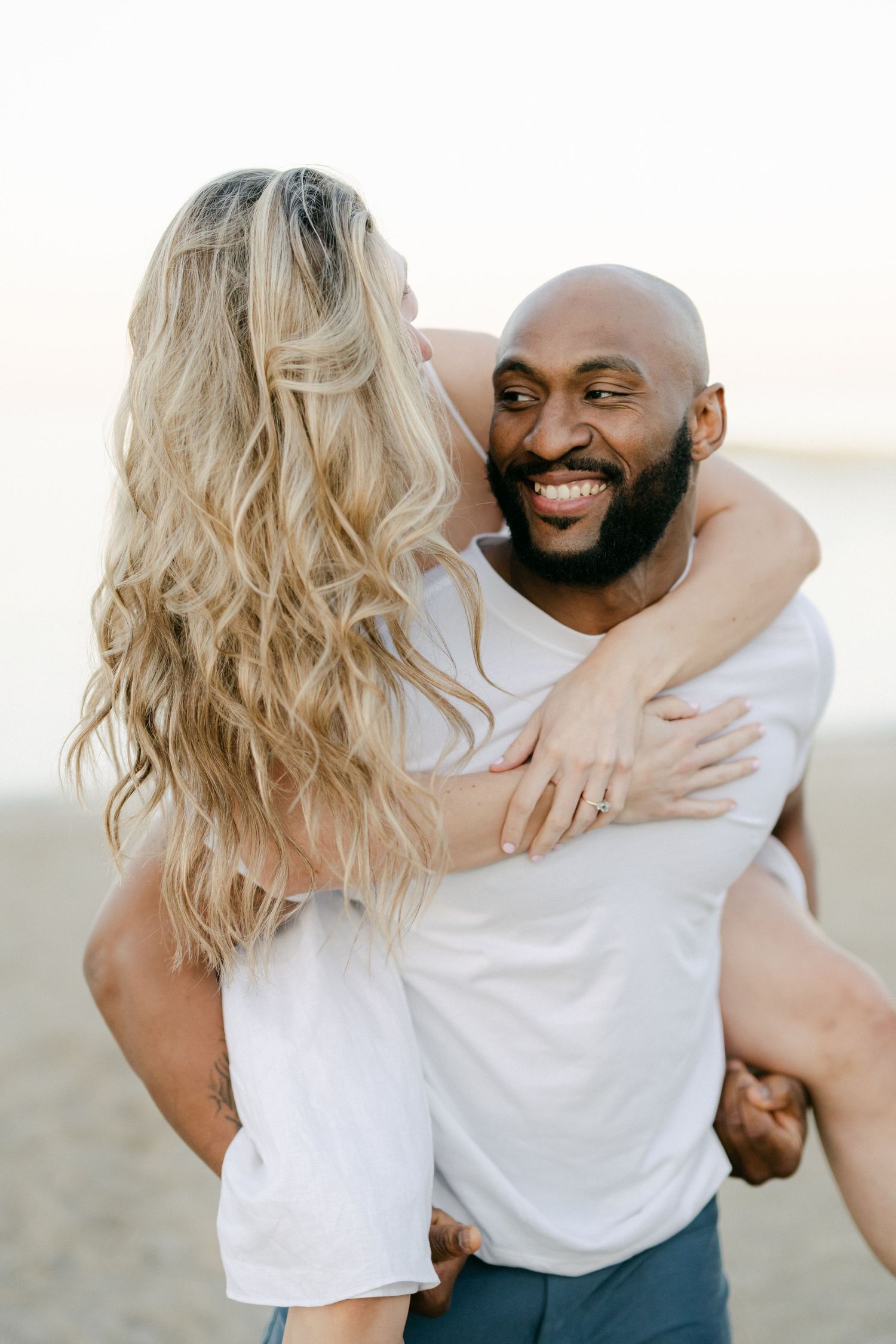 A couple lies on the beach laughing together while wearing casual white outfits during a romantic photoshoot. - Lake Forest
