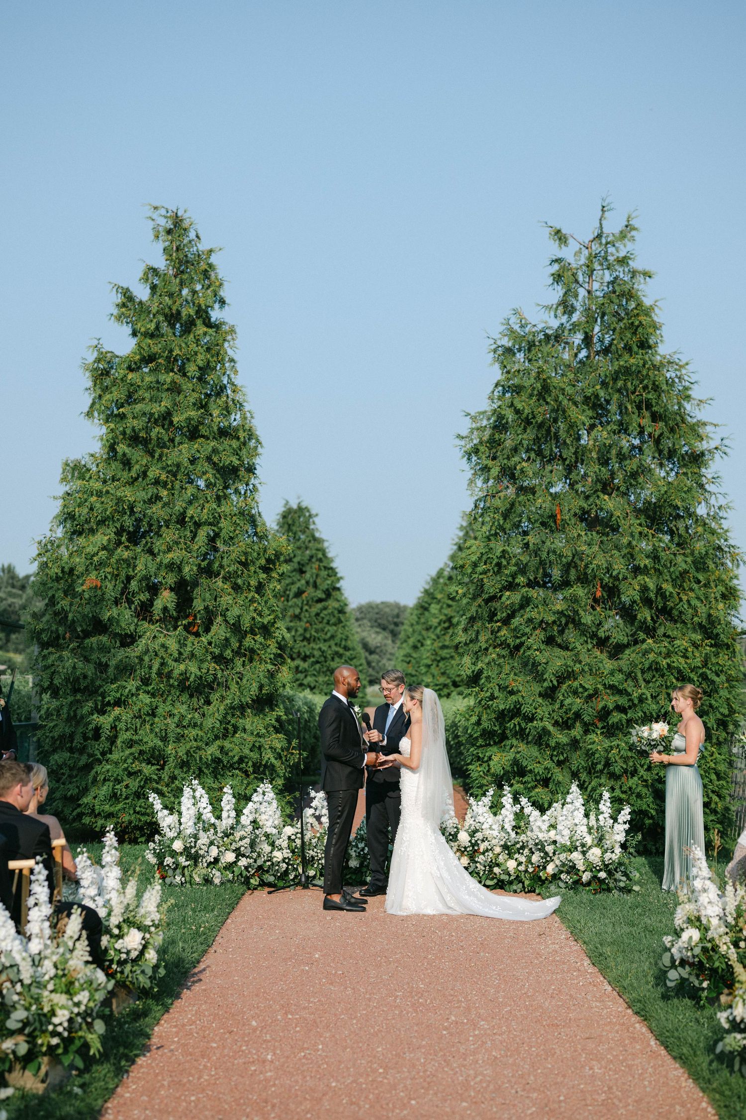 Bridesmaids in soft pink and sage green dresses gather together at an outdoor wedding ceremony with lush greenery. - Elawa Farm