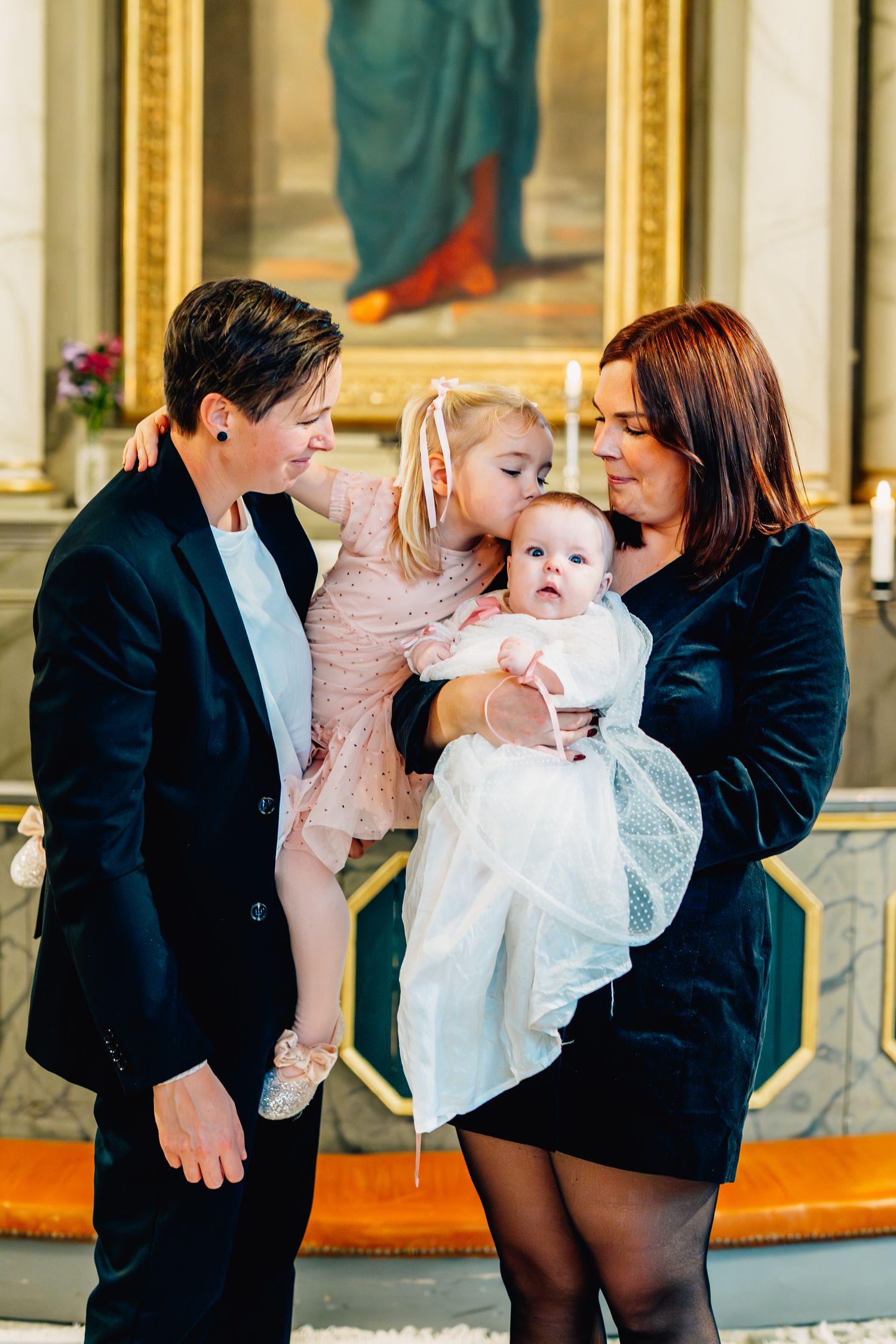 Ett familjeporträtt i kyrkan och storasyster pussar dopbarnet på huvudet.