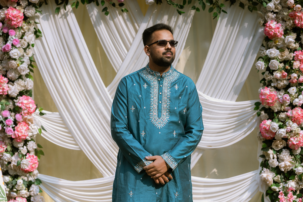 Person wearing a turquoise traditional kurta stands in front of white draped backdrop decorated with pink floral arrangements.