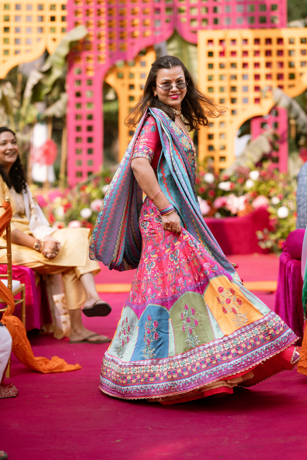 A dancer in a vibrant pink and blue lehenga twirls during a traditional Indian celebration on a red carpet.