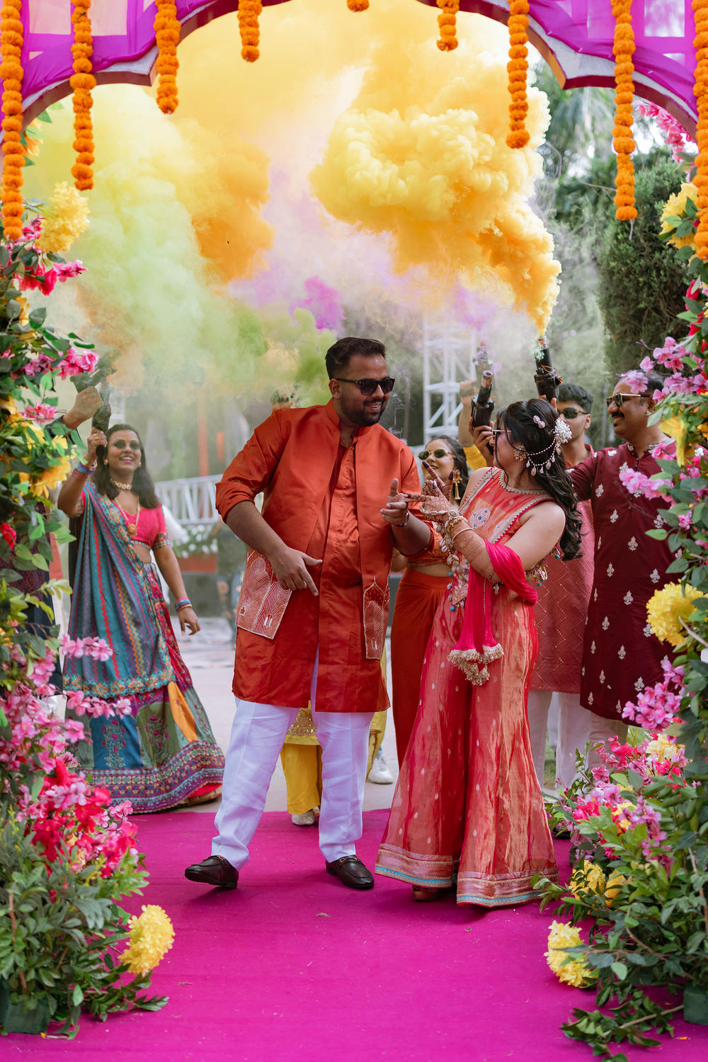 Colorful Indian wedding celebration with guests dancing amid vibrant yellow smoke and pink floral decorations.