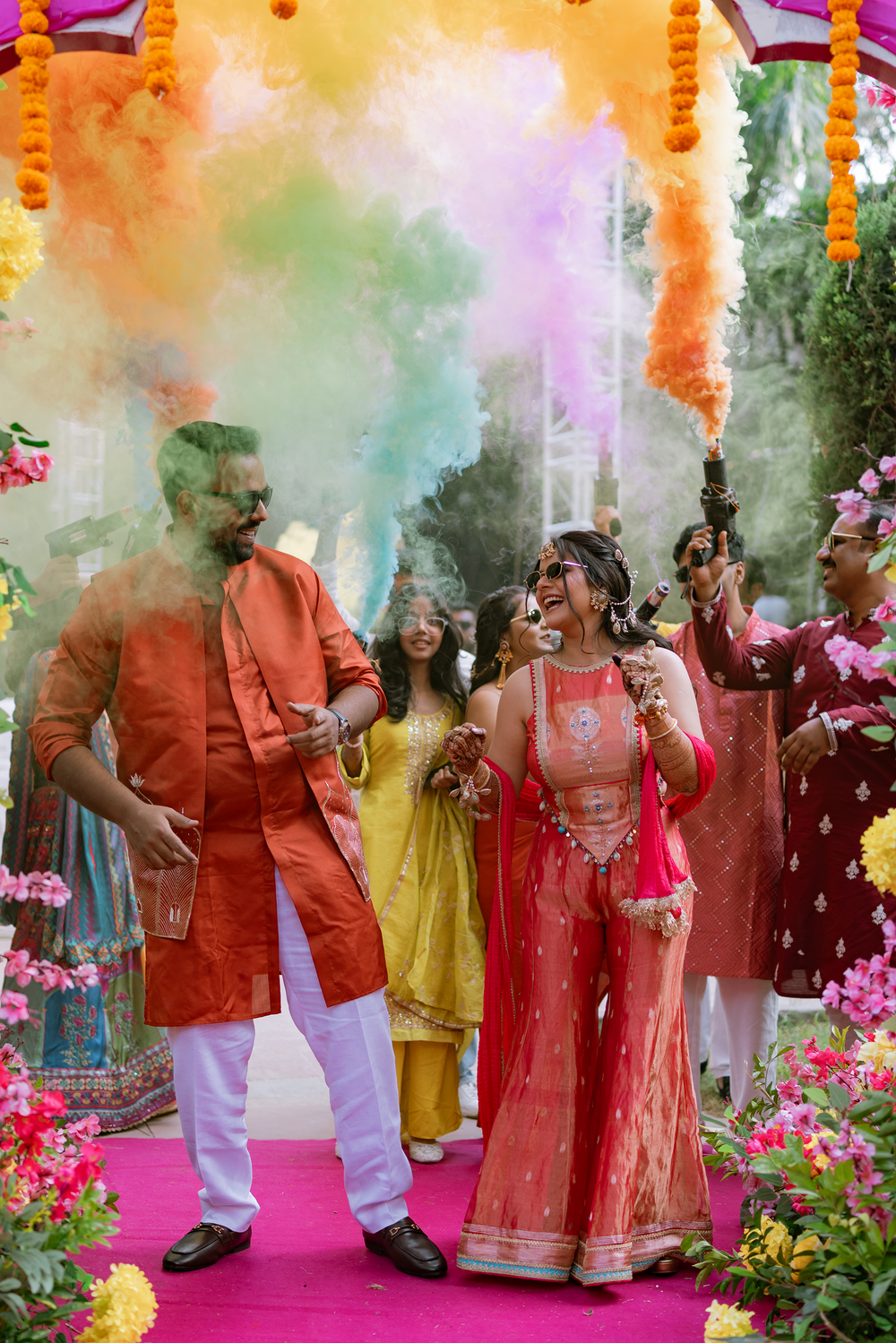 Traditional Indian wedding ceremony with guests in festive attire surrounded by marigold garlands and colored smoke effects.