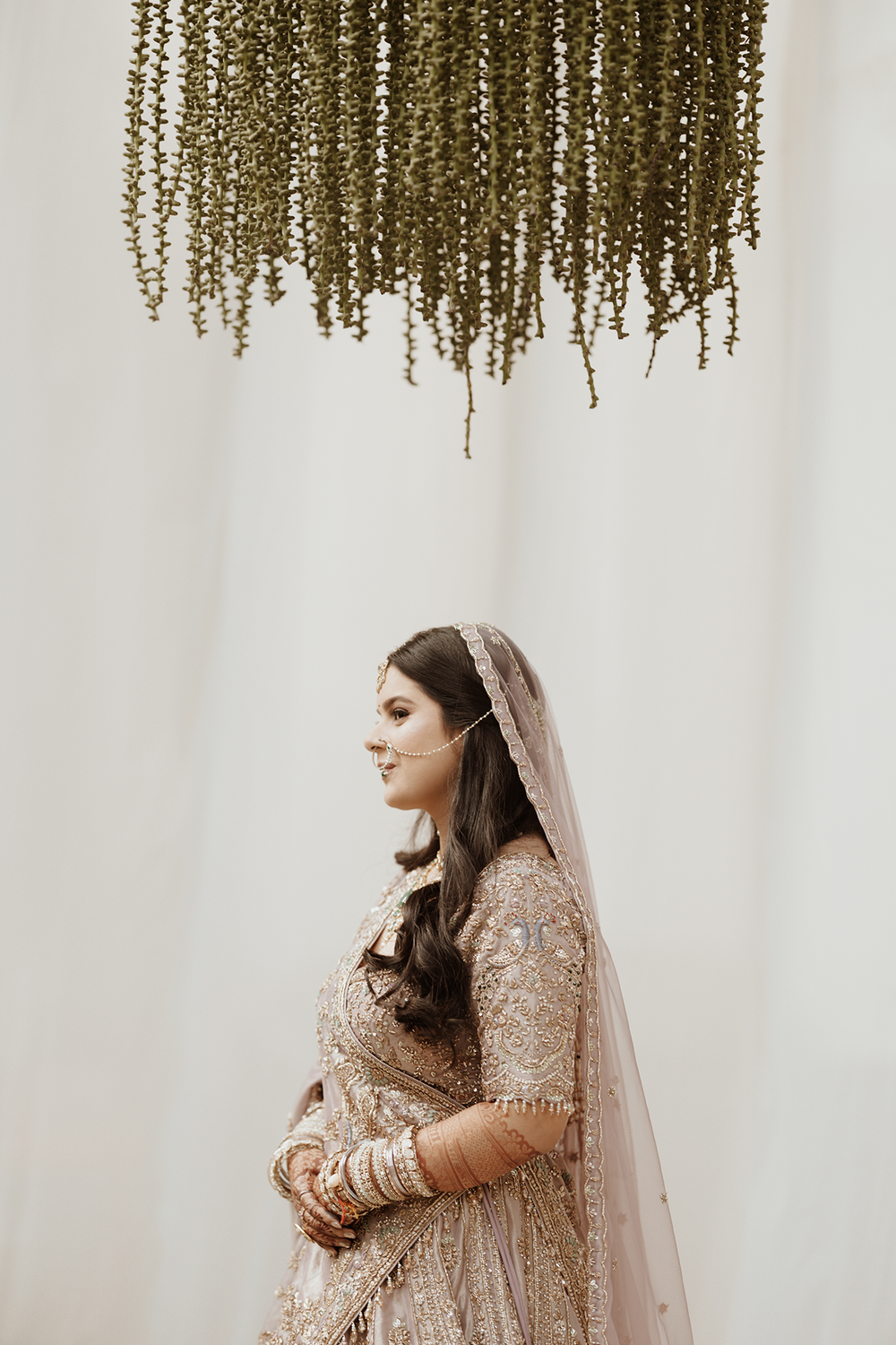 A bride in a beige embellished lehenga poses under hanging floral decor in an elegant wedding photoshoot.
