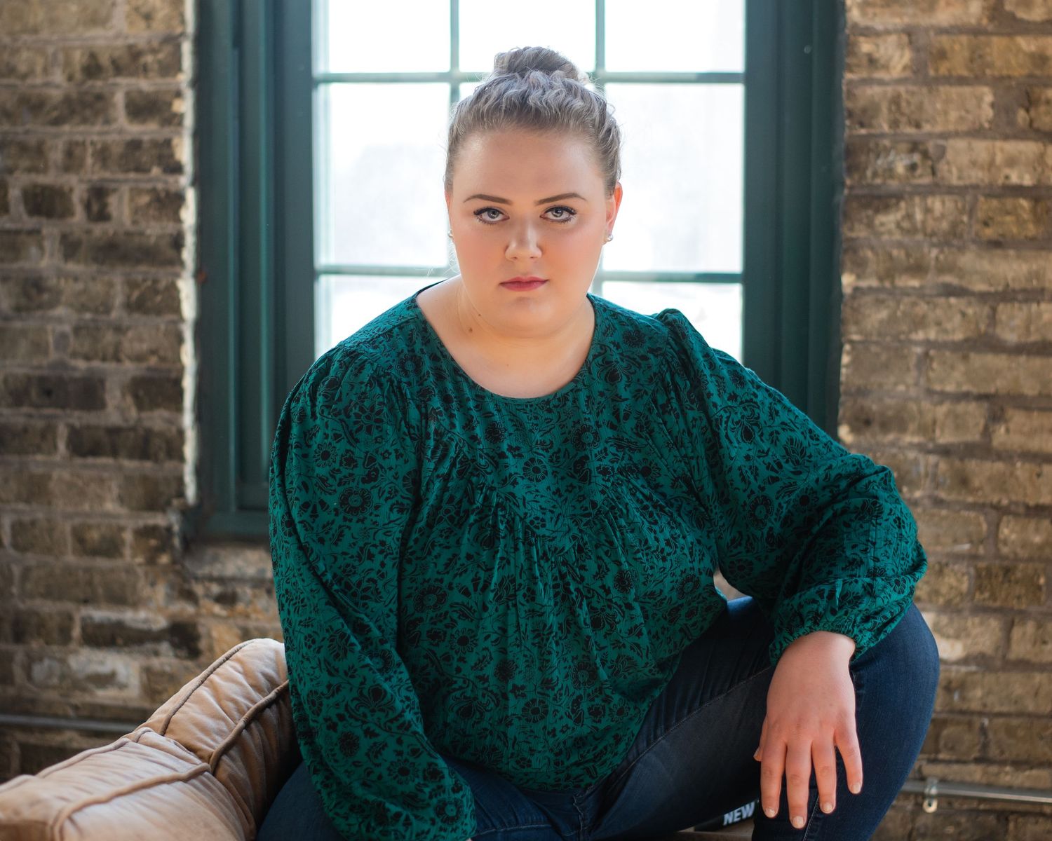 A person in an emerald green velvet dress poses against a brick wall with large windows.