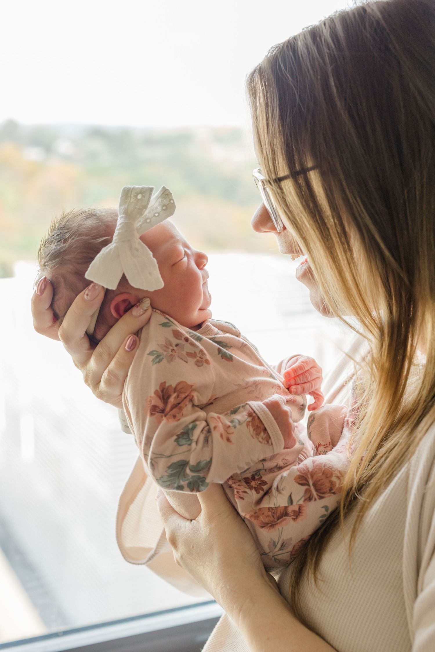 Parent holding and kissing a small baby wrapped in floral swaddle blanket near window during Fresh 48 session in Pittsburgh, PA.