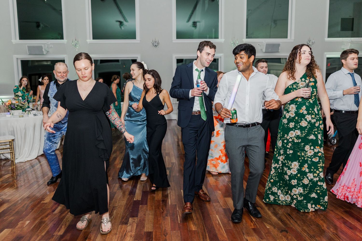 Wedding guests dance in formal attire on a wooden dance floor under bright lights.