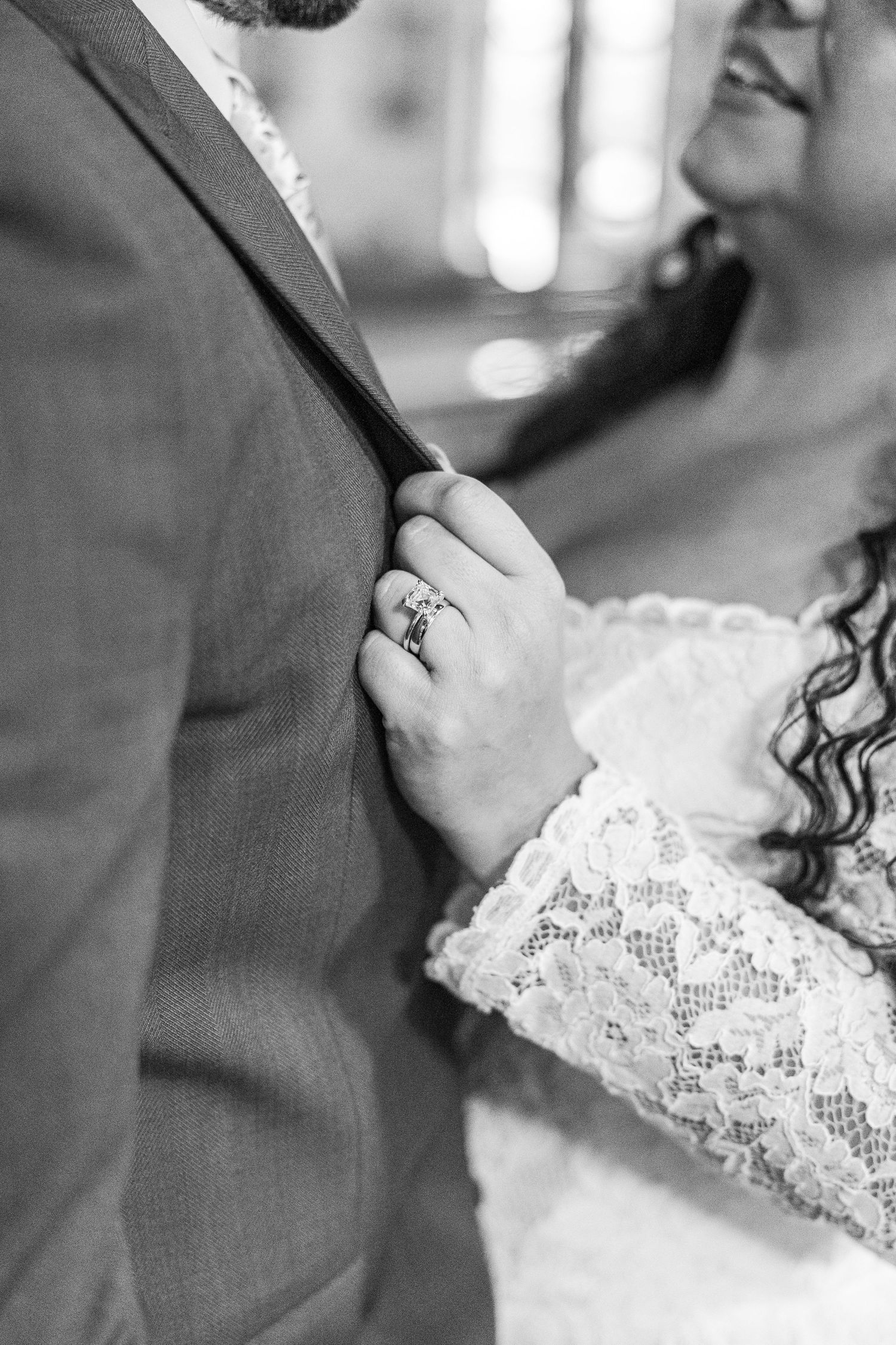 A close-up black and white photo of hands with a sparkling engagement ring against formal wedding attire.