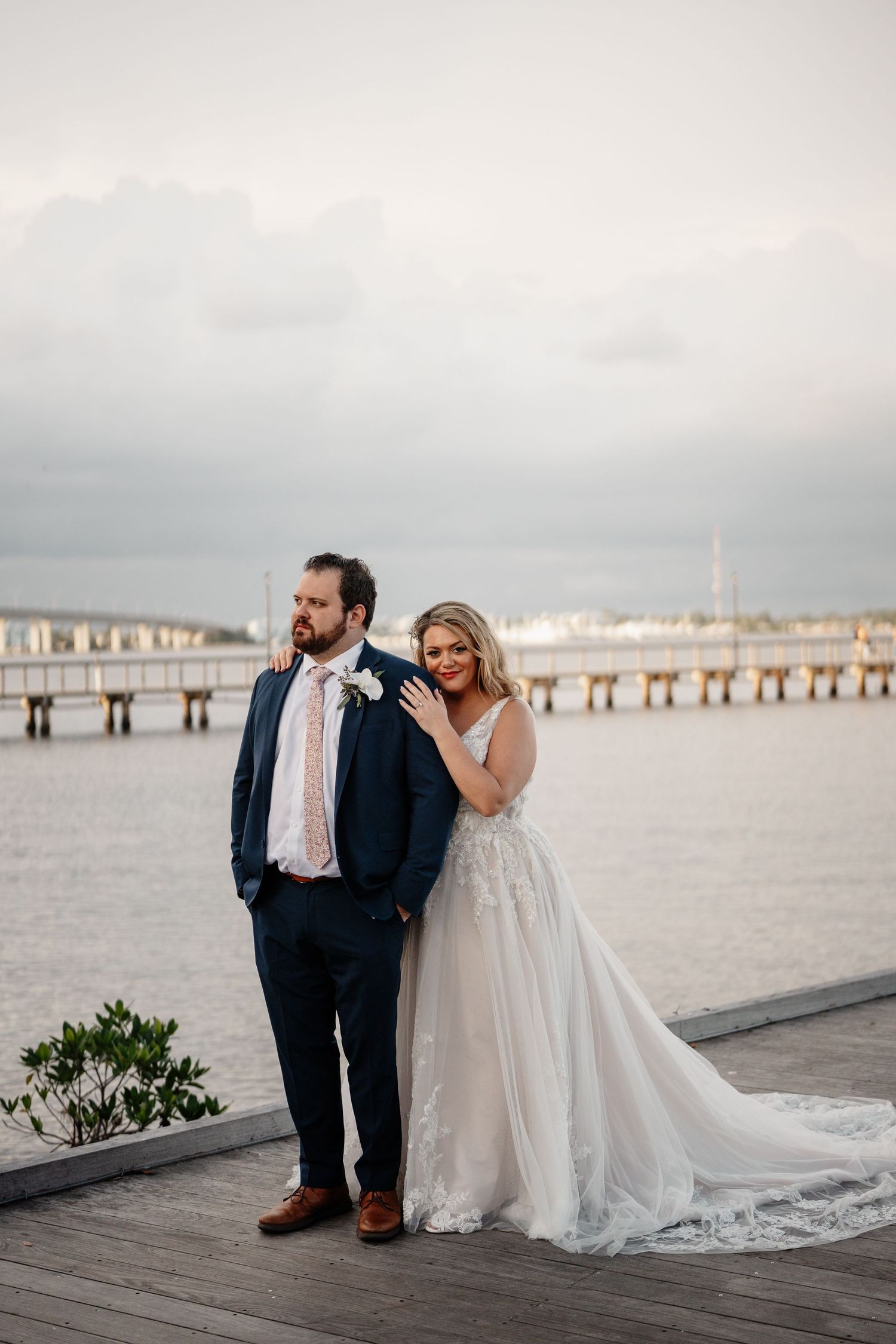 A couple in wedding attire stands together on a wooden pier overlooking the water at dusk.