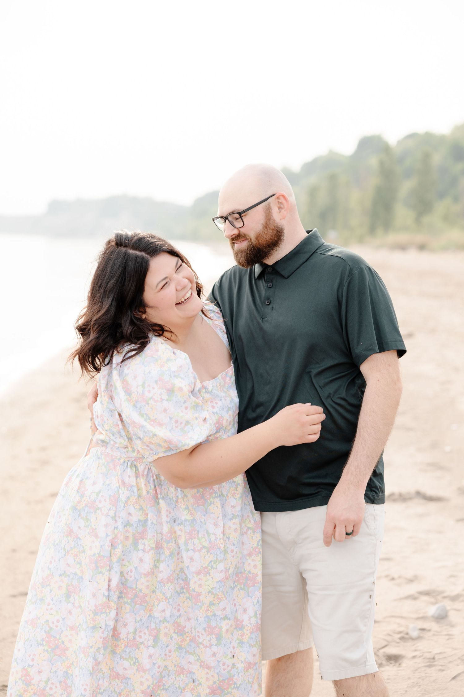 A couple shares a joyful moment together while standing on a sandy beach with trees in the background.