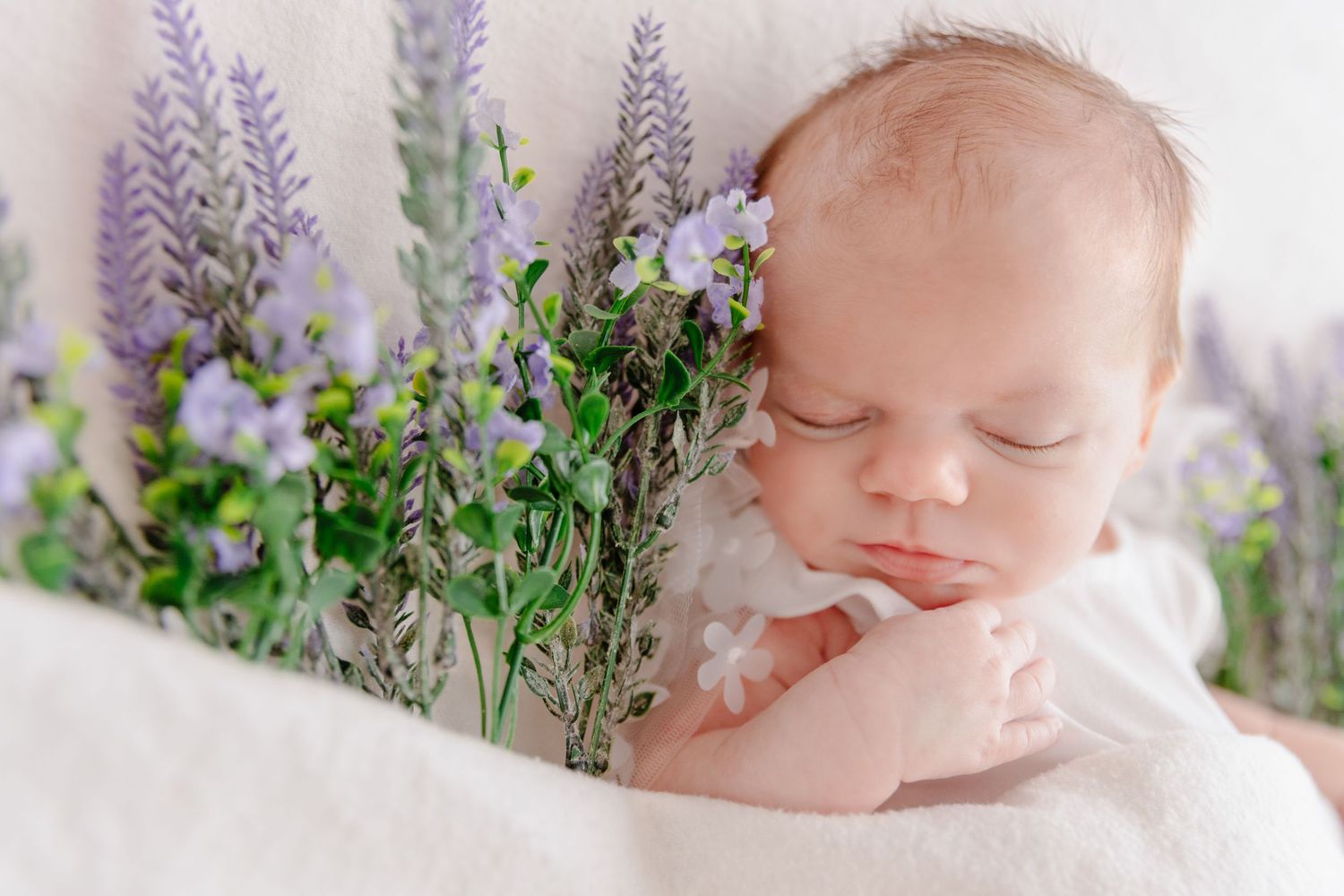 Sleeping baby in white wrap surrounded by delicate purple wildflowers and greenery in outdoor portrait setting.