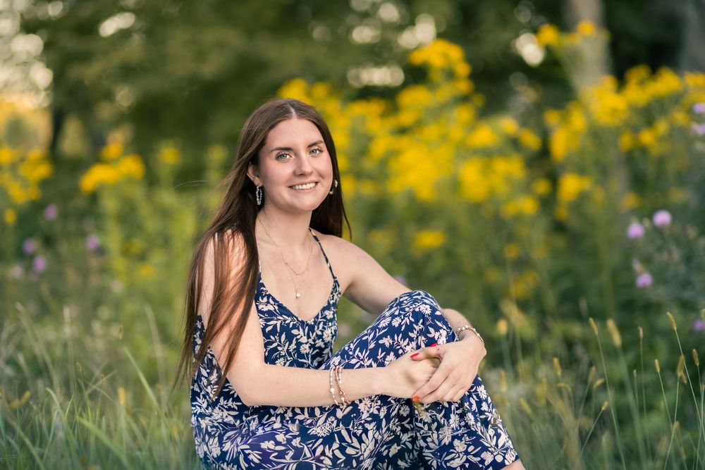 Woman in floral dress poses in a meadow with vibrant yellow wildflowers on a sunny summer day.