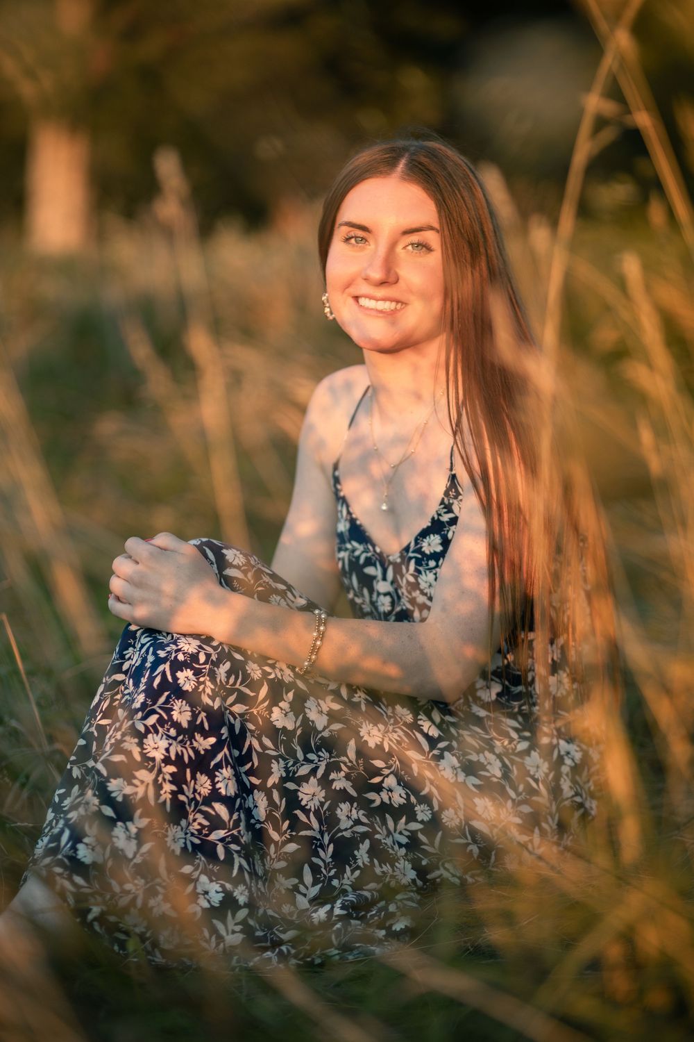 A person in a floral dress sits among tall grass during golden hour sunset lighting.