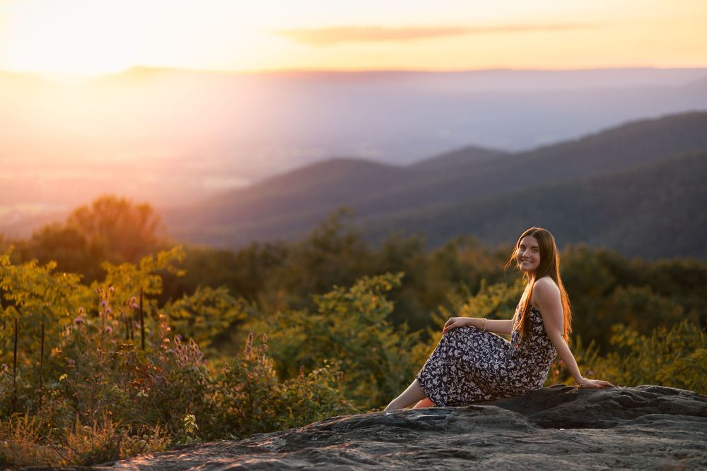A person sits on a rocky mountain overlook during golden sunset with rolling hills in the distance.