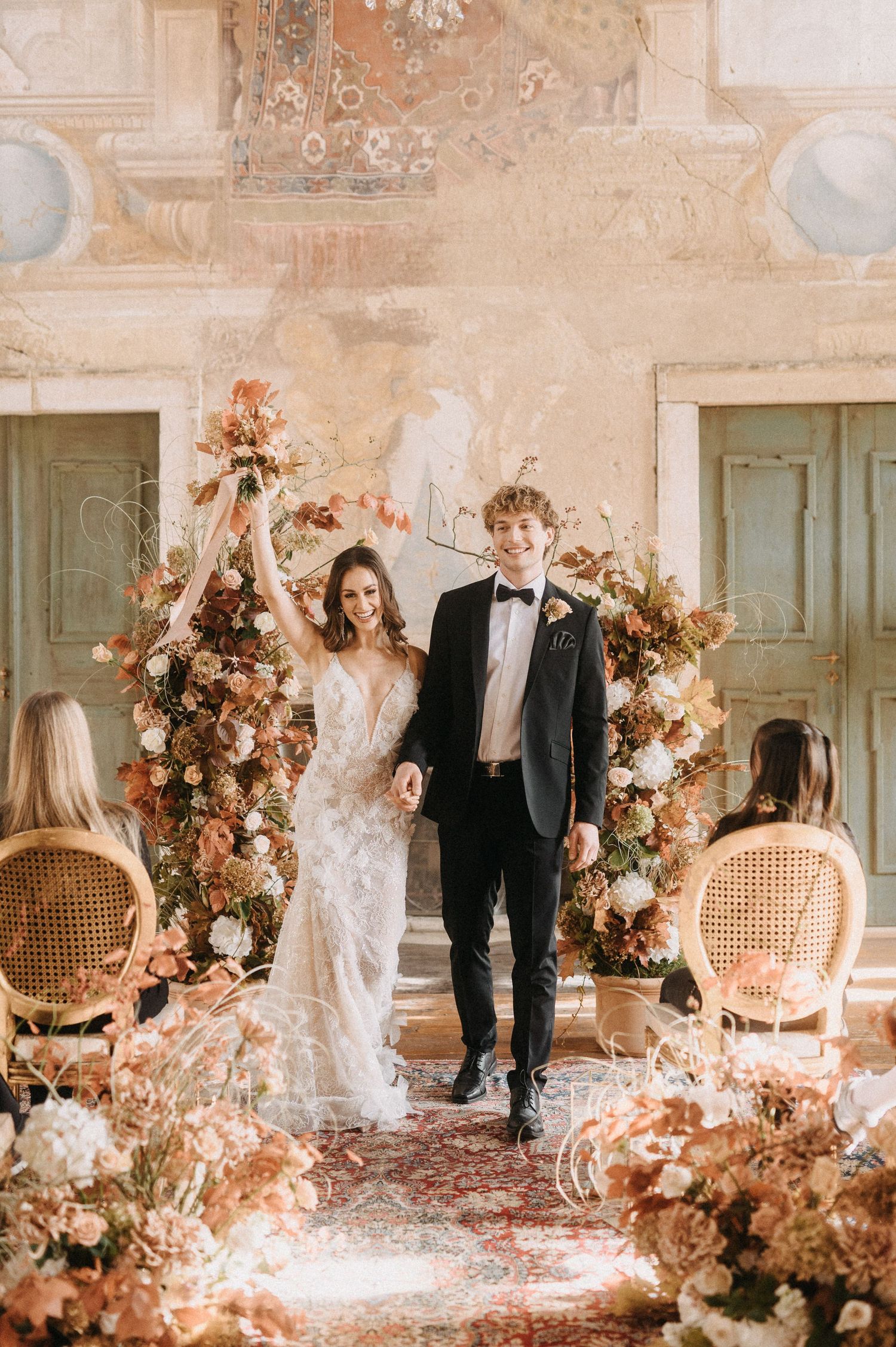 Newlyweds under a floral arch with peach-colored flowers in an elegant vintage setting.